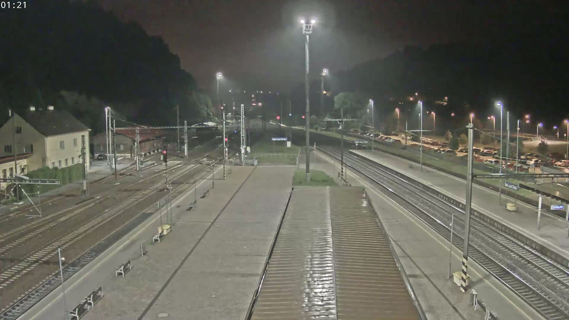 A nighttime view of a train station with multiple tracks, platforms, and a building, surrounded by dark hills and some parked cars under artificial lighting.