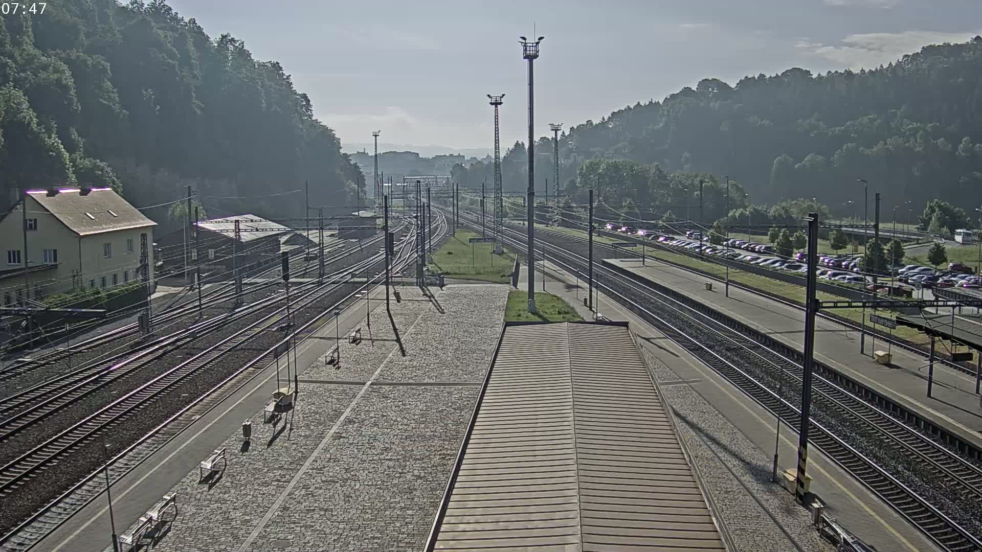A train station with multiple tracks, a large parking lot, and a building, is situated against a backdrop of a hill covered in green trees under a clear sky.