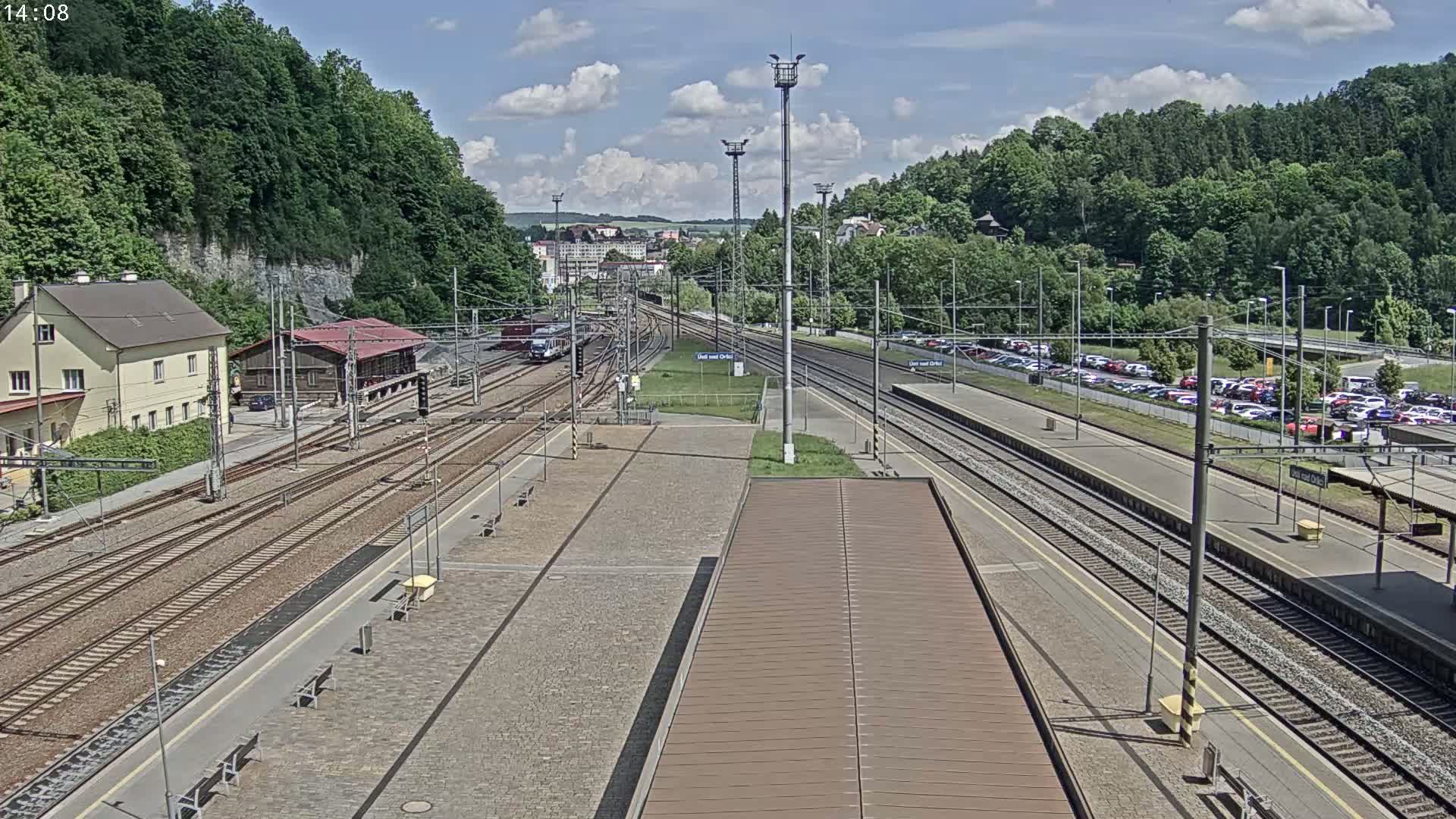 A train station with multiple train tracks, a building, and a parking lot, under partly cloudy skies.