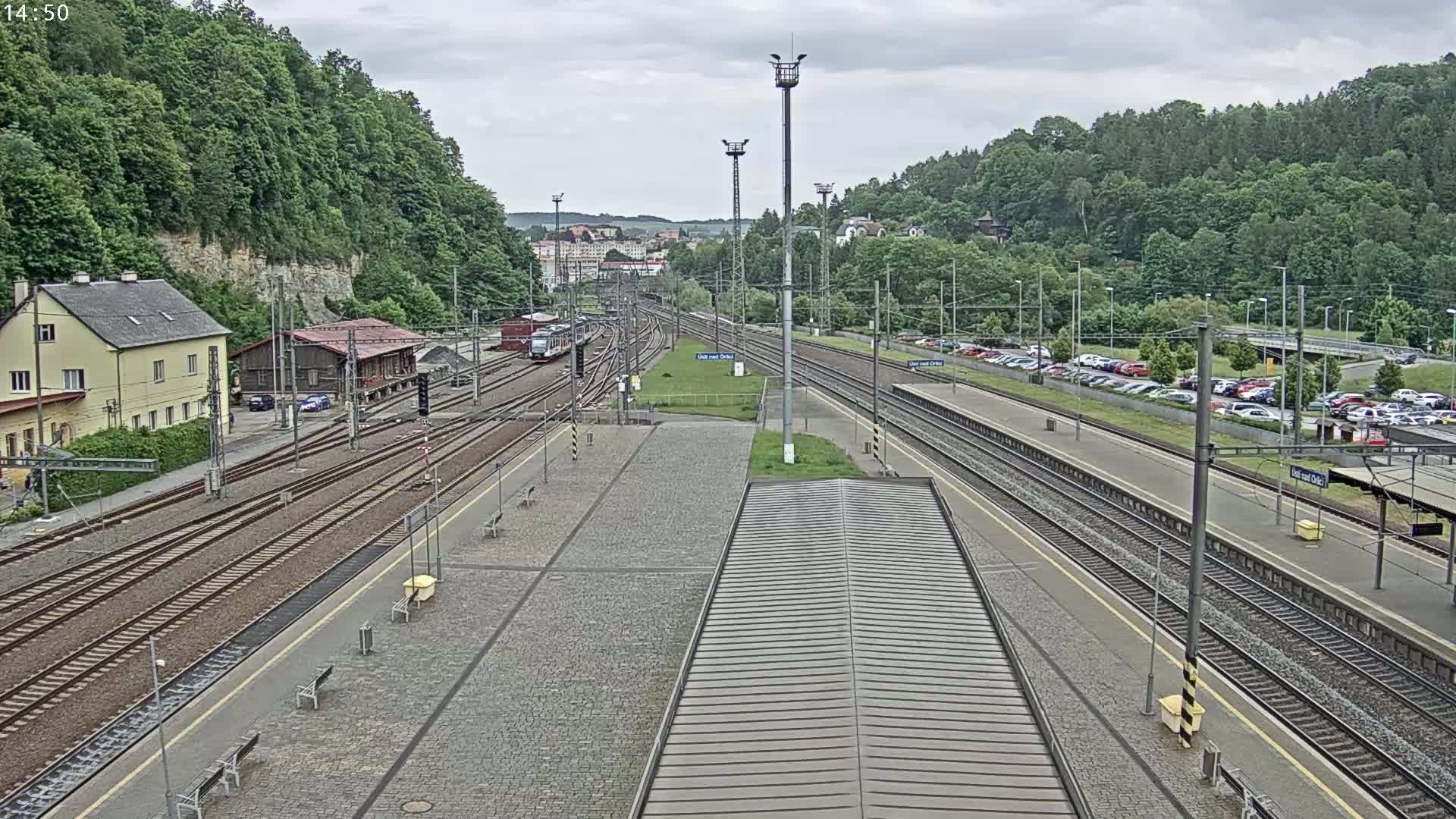 A train station with multiple train tracks, a parking lot full of cars, and a building is seen on an overcast day.
