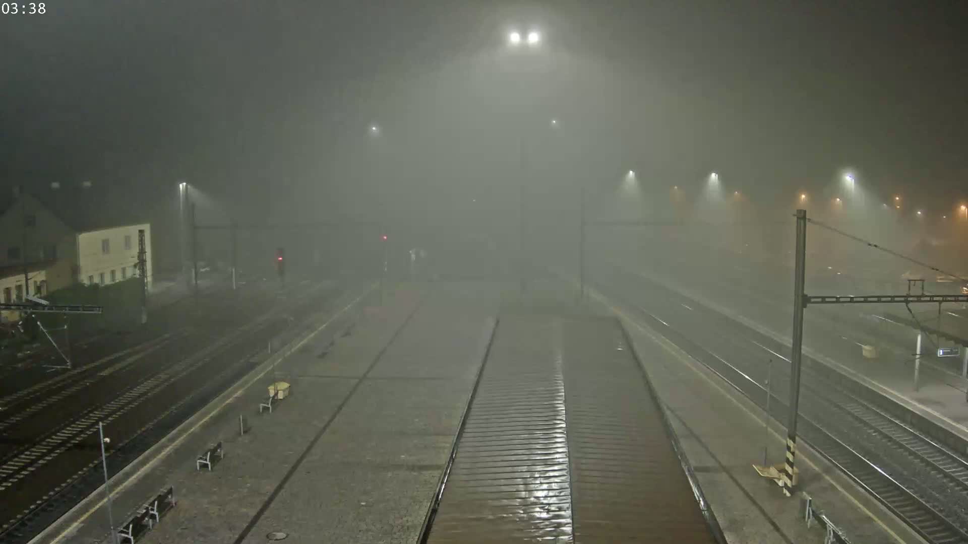 A train station platform is shown at night in heavy fog and rain.