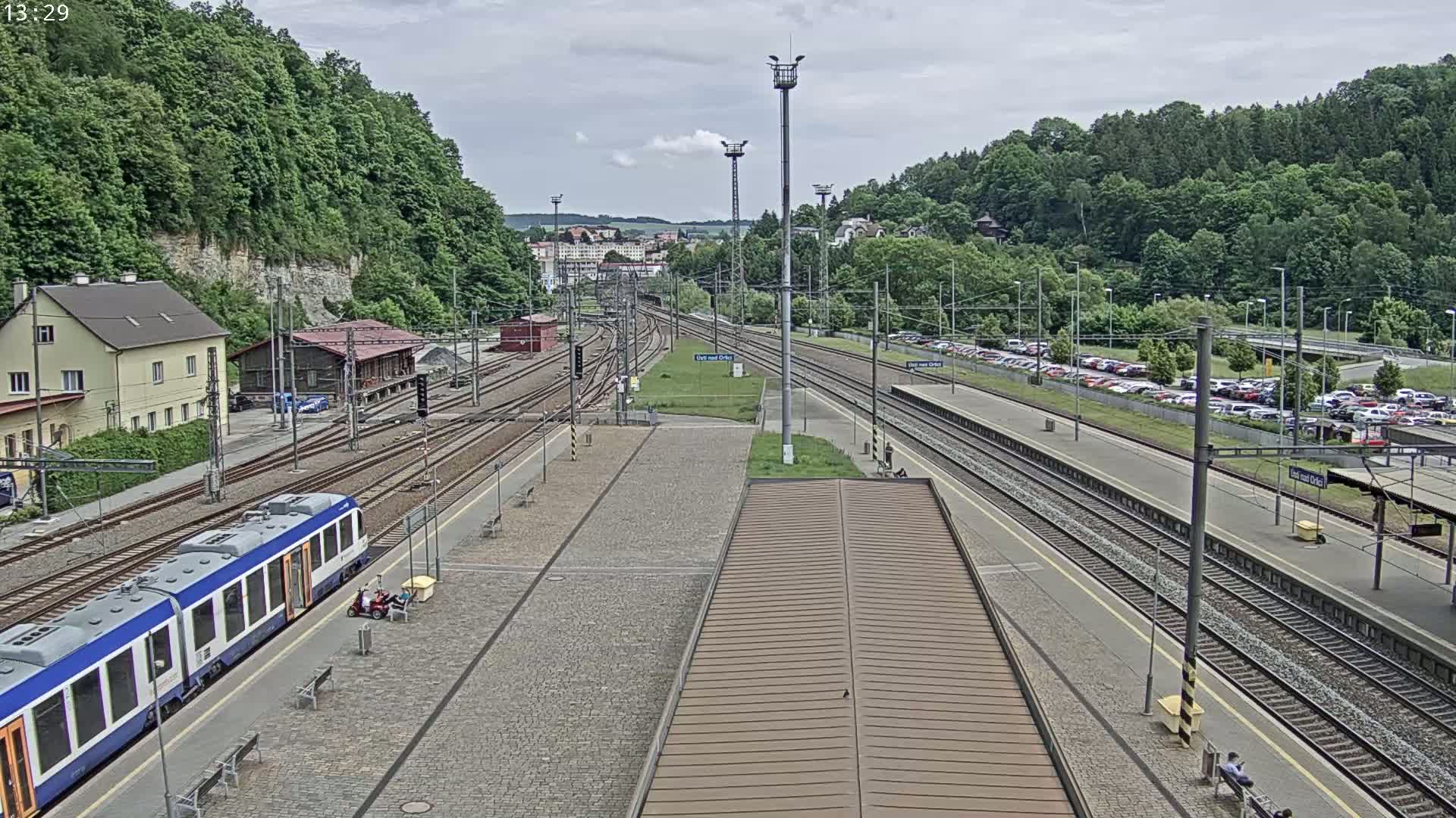 A train station with multiple train tracks, a blue and white train, and a large parking lot is visible on an overcast day.