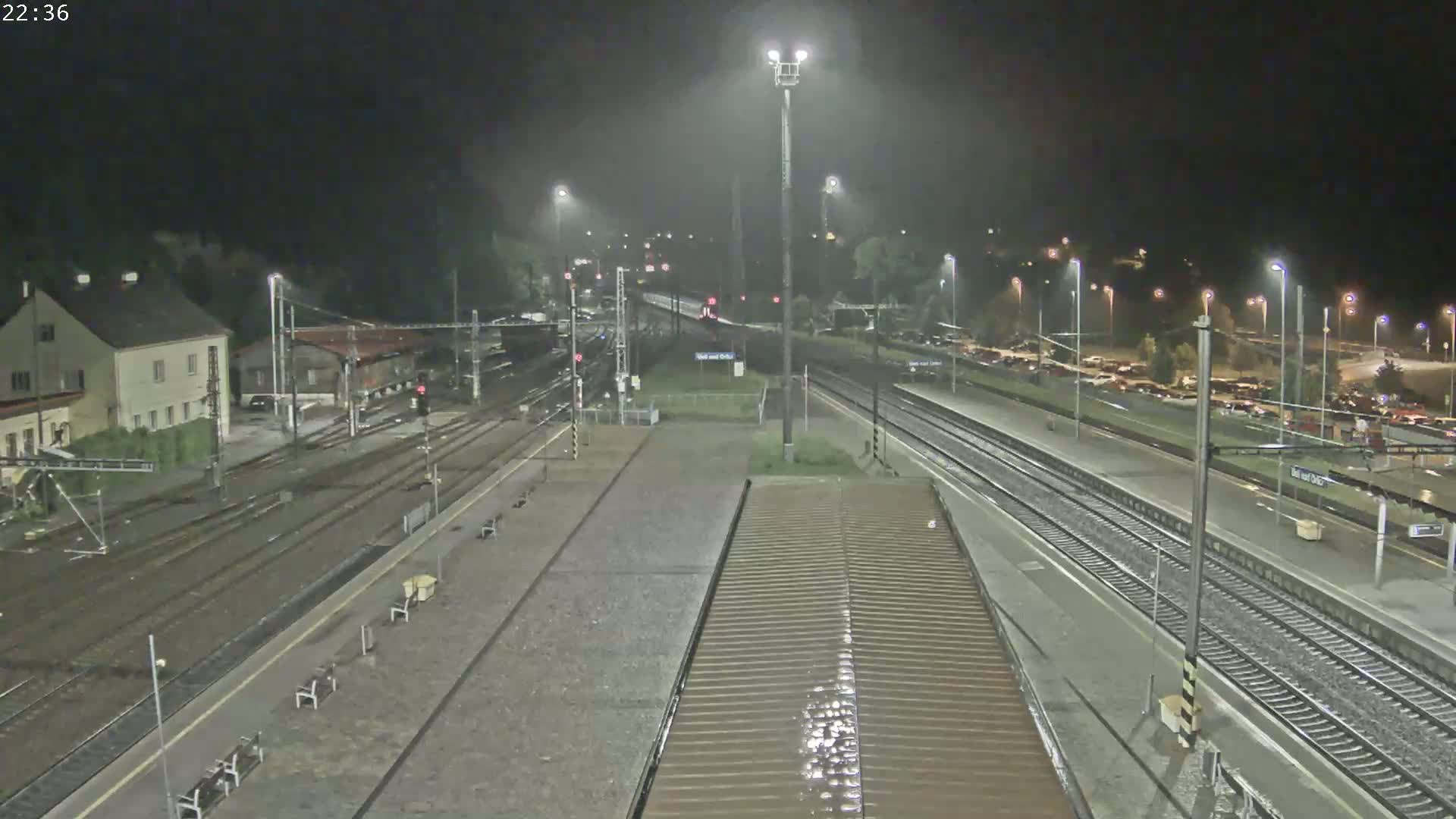 A nighttime view of a rain-slicked train station with multiple tracks, platforms, and a building in the background.