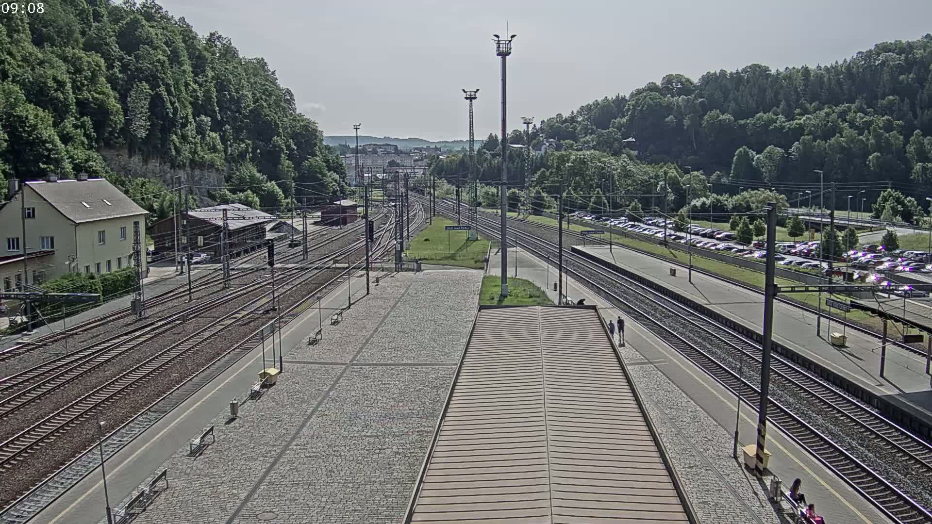 A high-angle view of a train station on a sunny day, showing multiple train tracks, platforms, a parking lot filled with cars, and a surrounding landscape of green hills and trees.