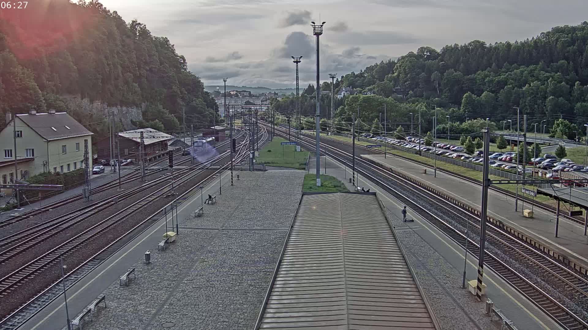 A mostly cloudy day overlooks a train station with multiple tracks, a parking lot full of cars, and a building situated on a hillside covered in green trees.
