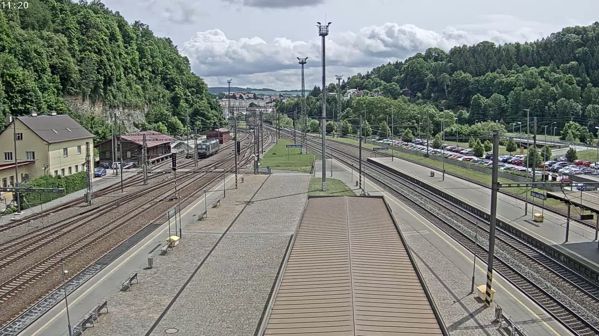 A mostly sunny day at a train station with multiple tracks, a large parking lot, and a building nestled against a verdant hillside.