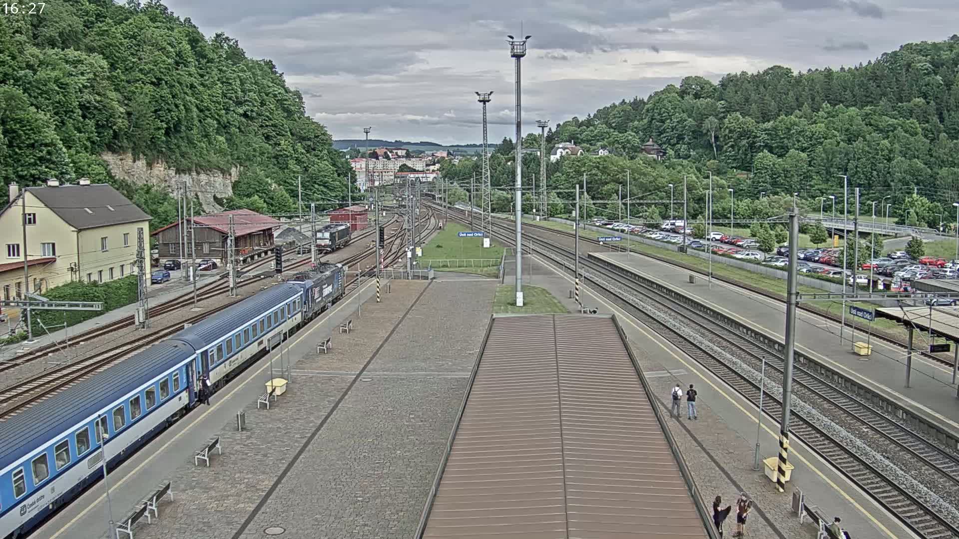 A train station with a passenger train at a platform, multiple train tracks, a large parking lot, and surrounding green hills under an overcast sky.