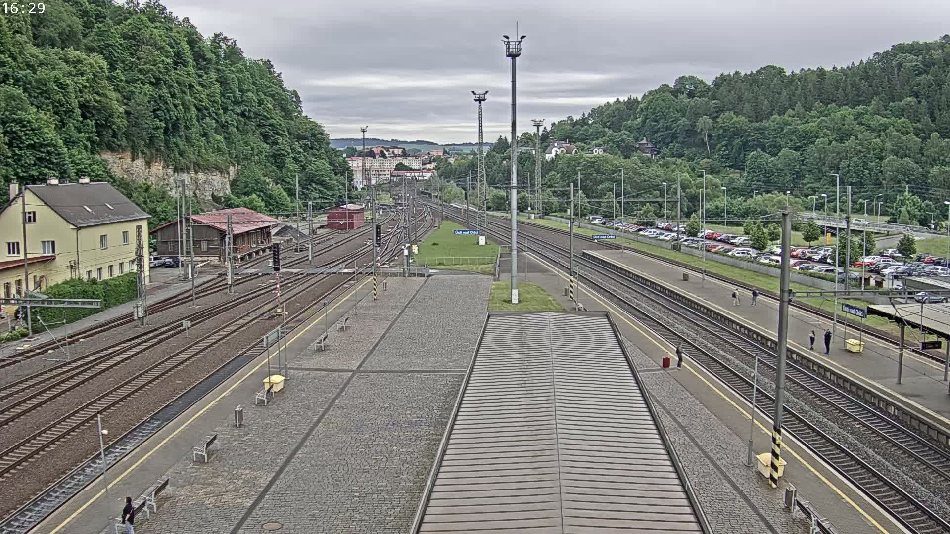 An overcast day reveals a train station with multiple tracks, a large parking lot, and buildings surrounded by lush green hills.