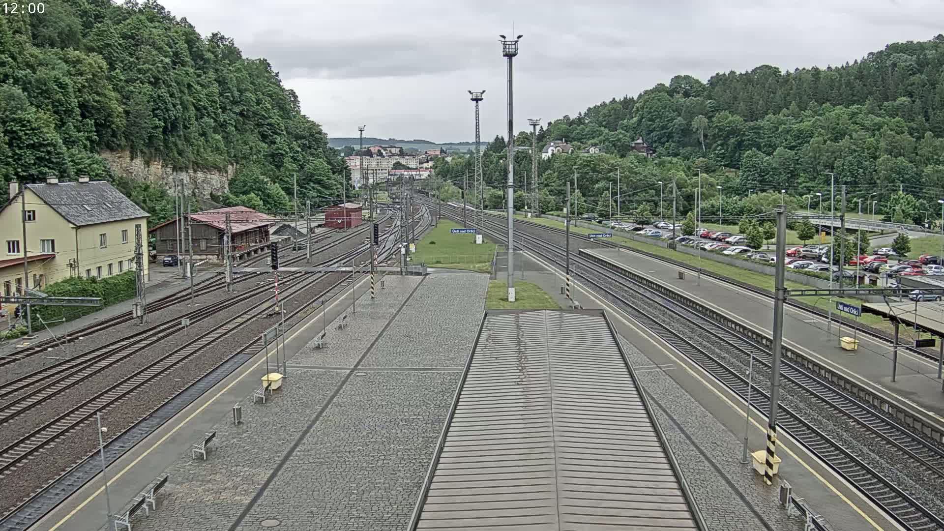 A high-angle view of a train station on an overcast day, showing multiple train tracks, a platform, a building, and a parking lot filled with cars, all surrounded by lush green hills.