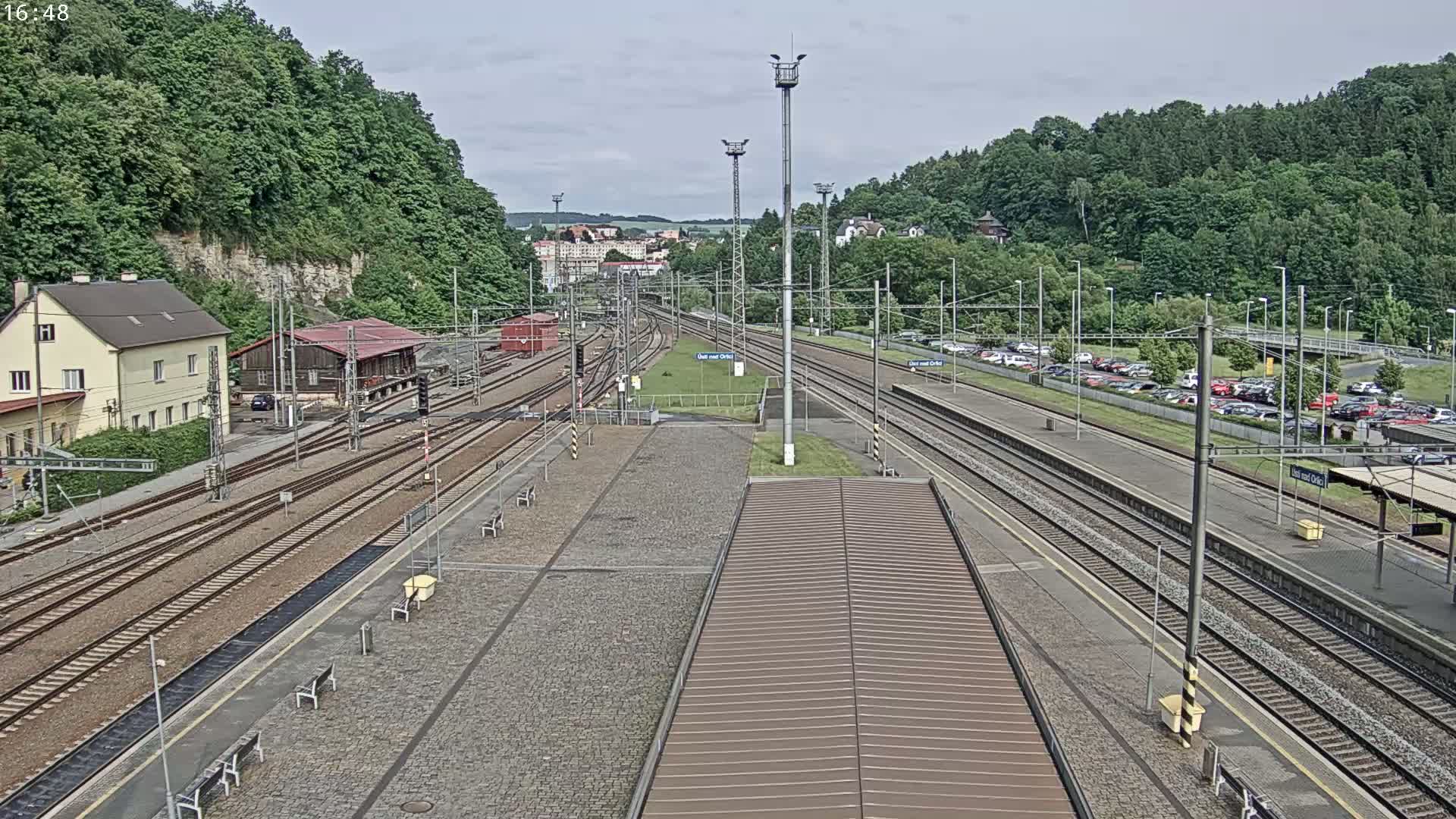 A train station with multiple train tracks, a parking lot, and buildings surrounded by green hills on an overcast day.