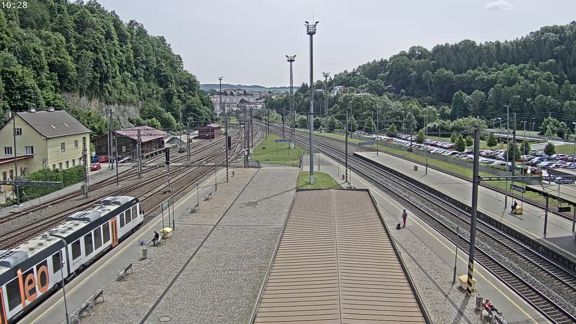 A train station with multiple train tracks, a passenger train, a large parking lot, and a hillside covered in lush green trees under a partly sunny sky.