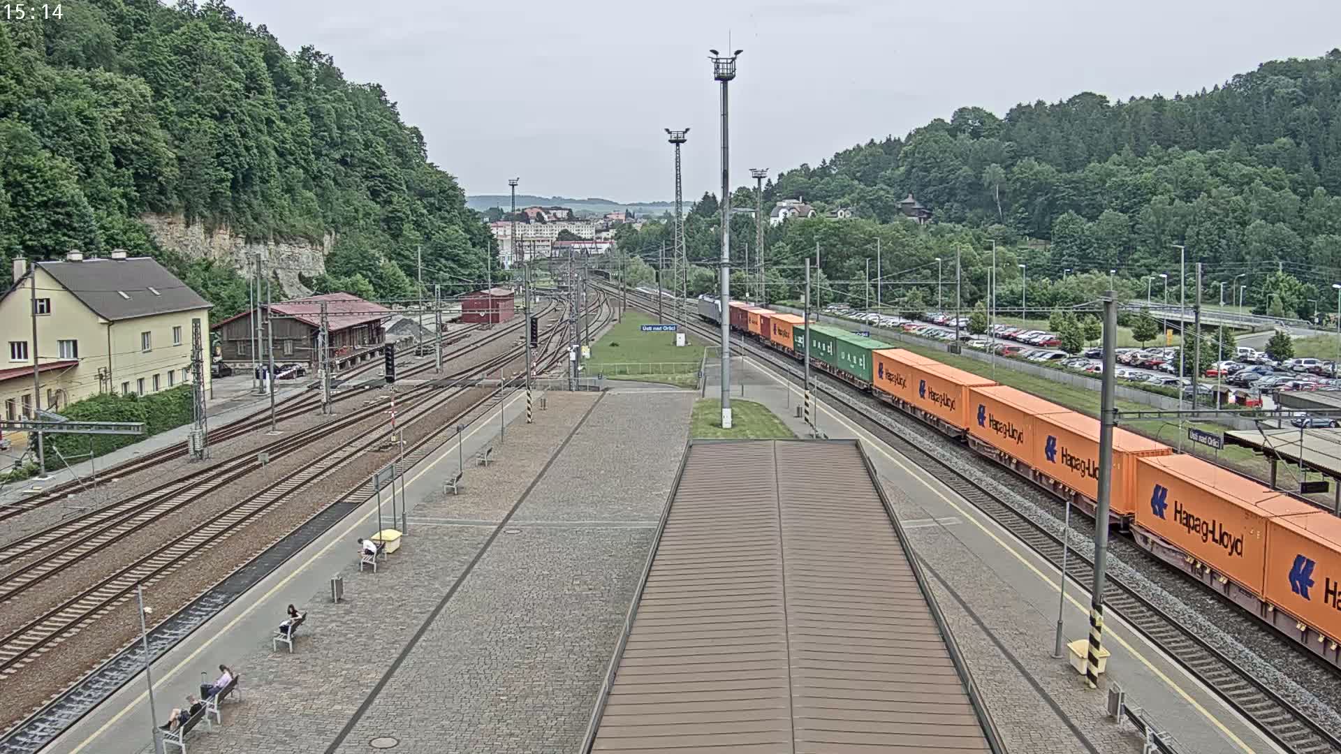 A freight train carrying orange and green containers passes through a train station on an overcast day, alongside a paved platform with several people sitting on benches, with a hillside covered in lush green trees in the background.