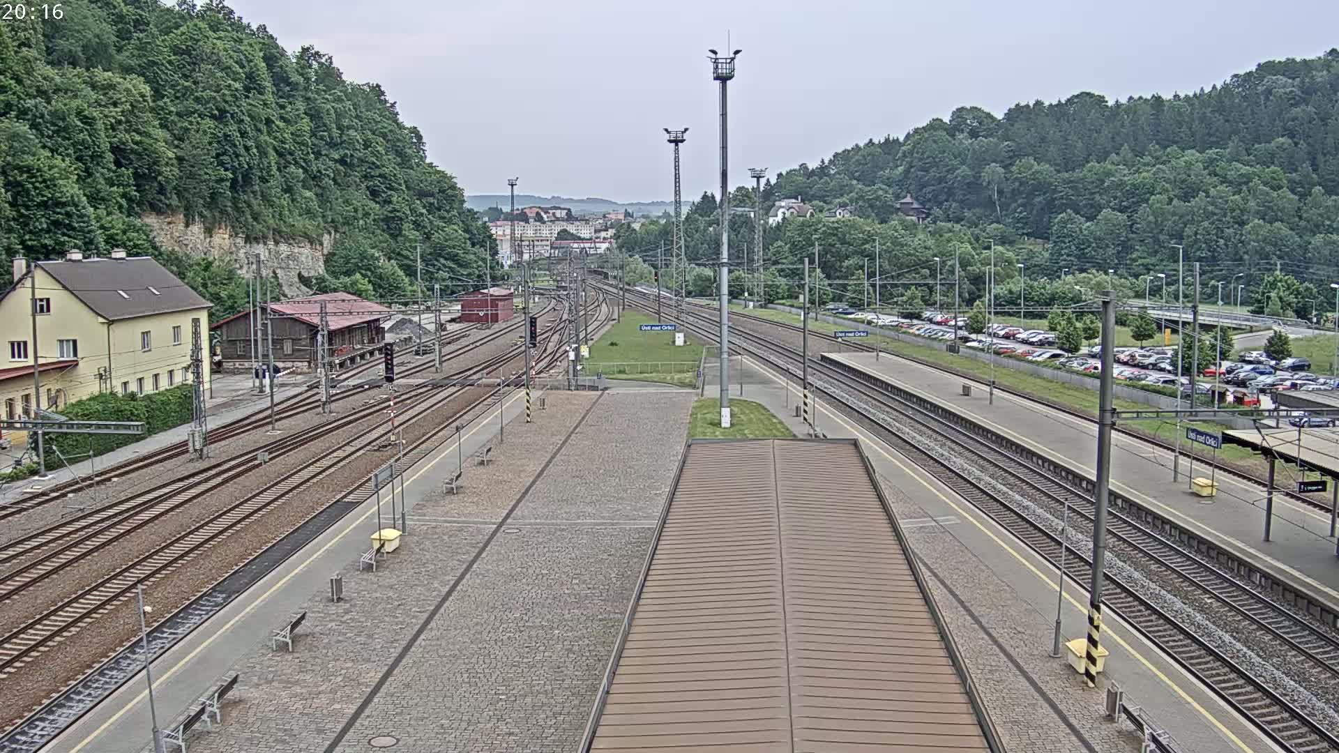 A train station with multiple tracks, a large parking lot, and buildings surrounded by a lush green hillside under an overcast sky.