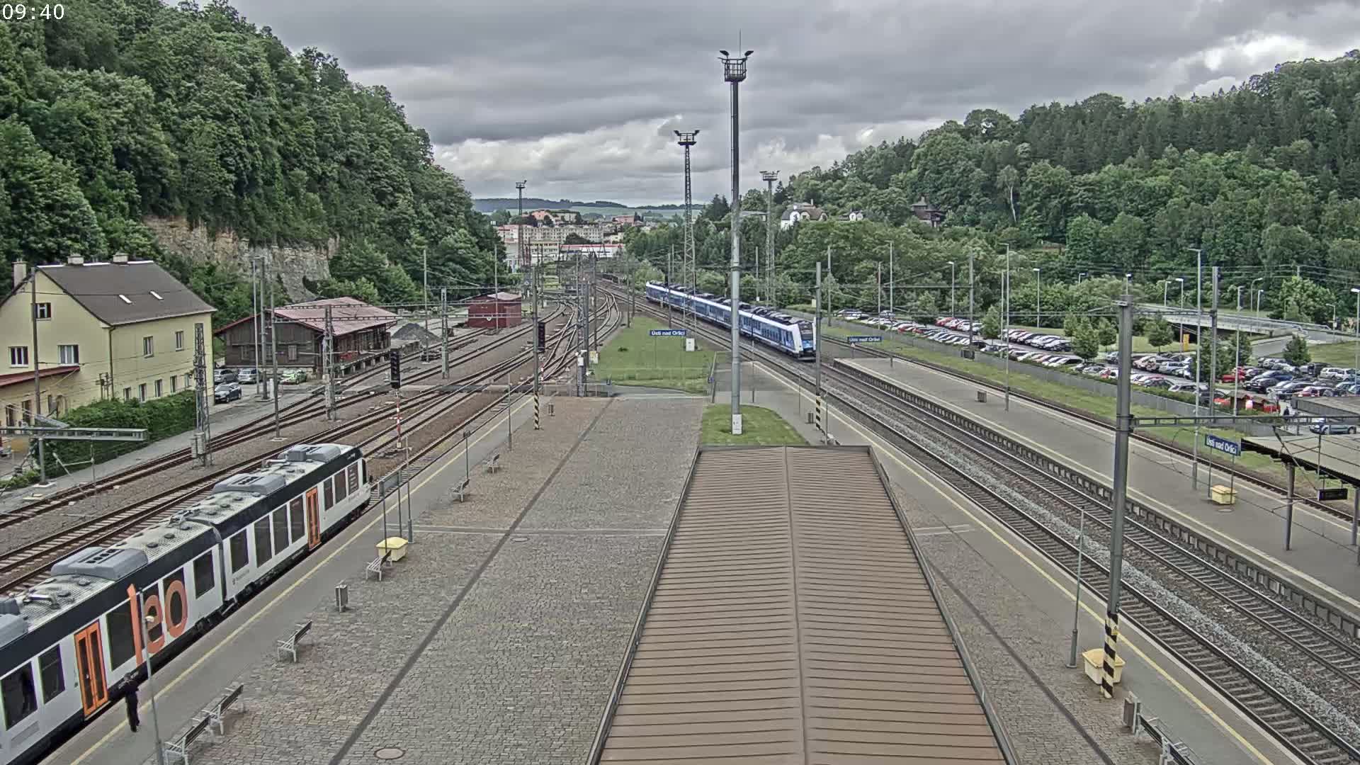 A train station with multiple train tracks, a long train departing, and a smaller train arriving, surrounded by green hills under an overcast sky.