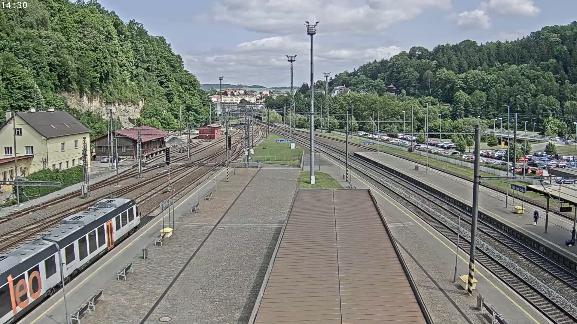 A train station with multiple tracks, a train on a platform, buildings, and a large parking lot is seen under a partly cloudy sky.