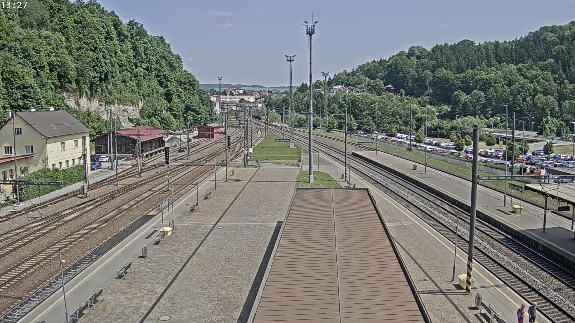 A train station with multiple tracks and a large parking lot is visible on a sunny day, surrounded by lush green hills.