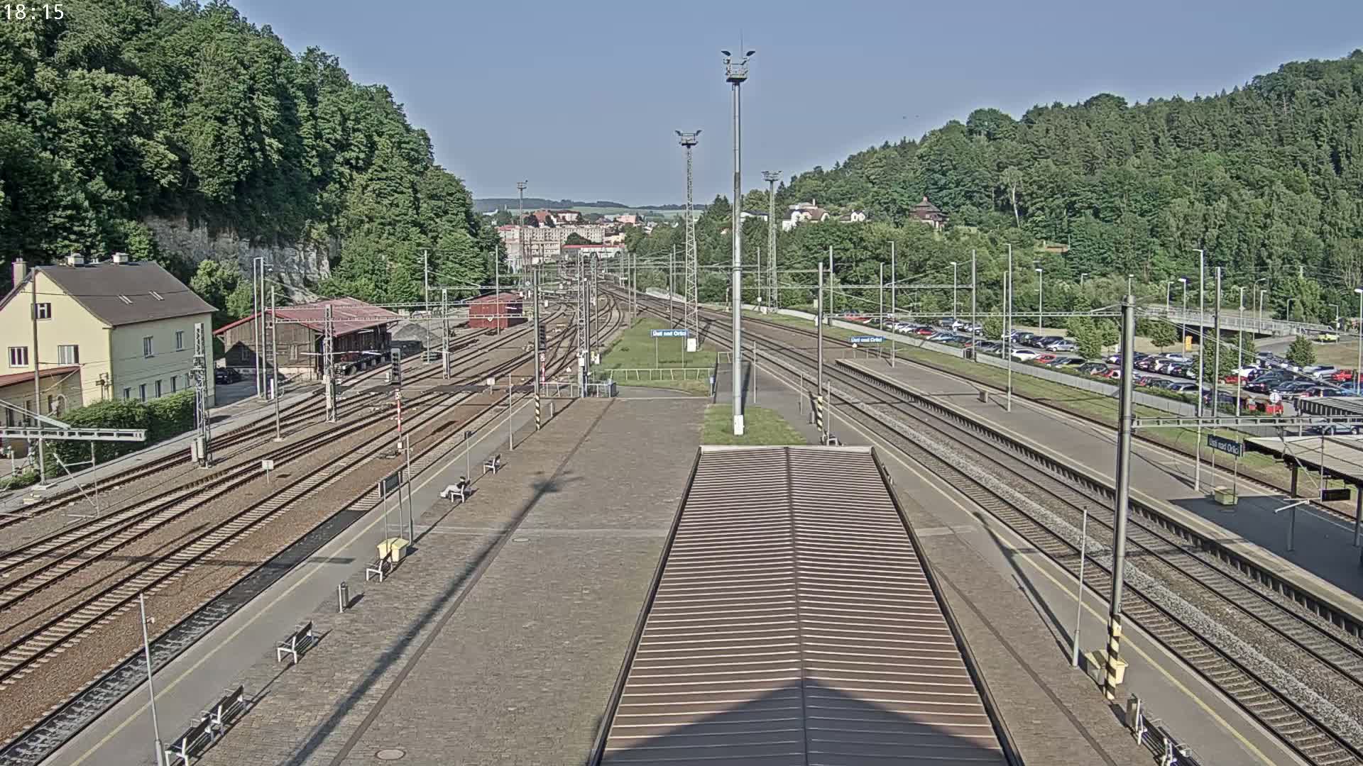 A high-angle view of a train station on a sunny day, showing multiple train tracks, platforms, a parking lot, and surrounding green hills.