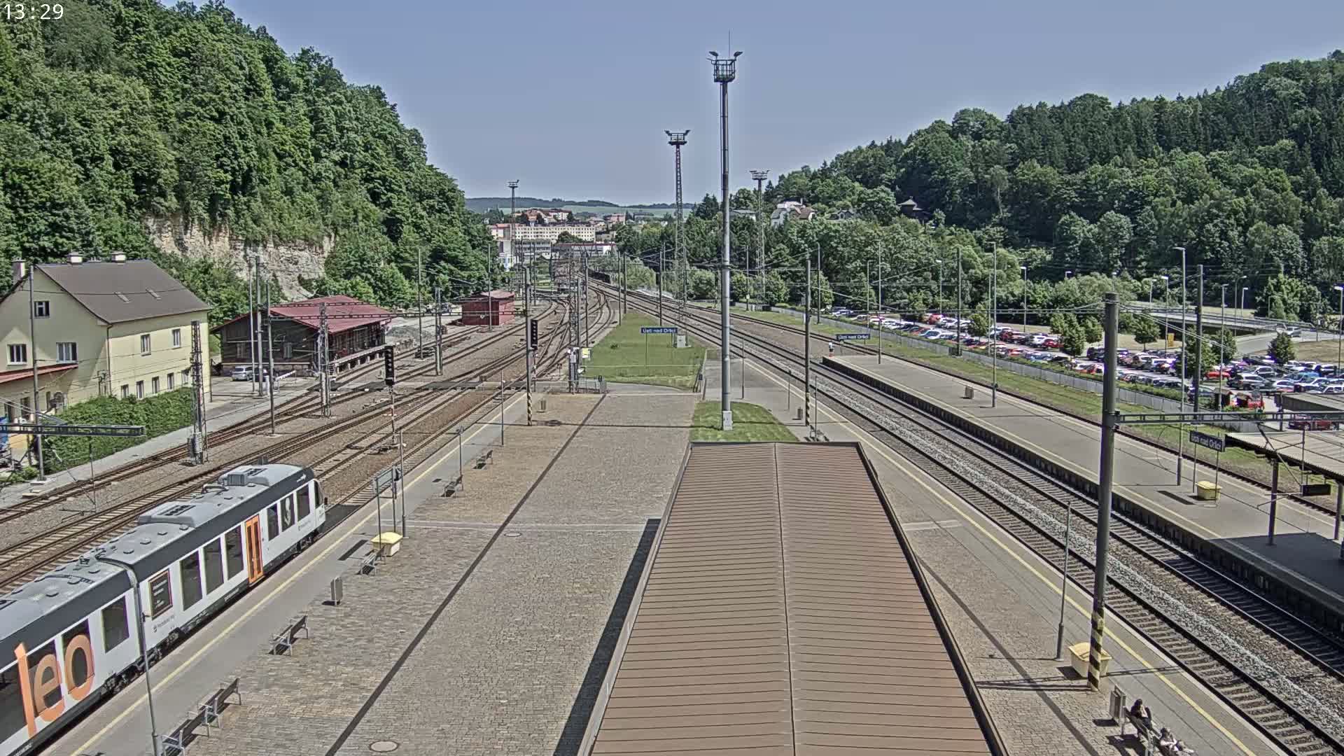 A train station with multiple train tracks, a passenger train, and a large parking lot is seen on a sunny day, surrounded by lush green hills.