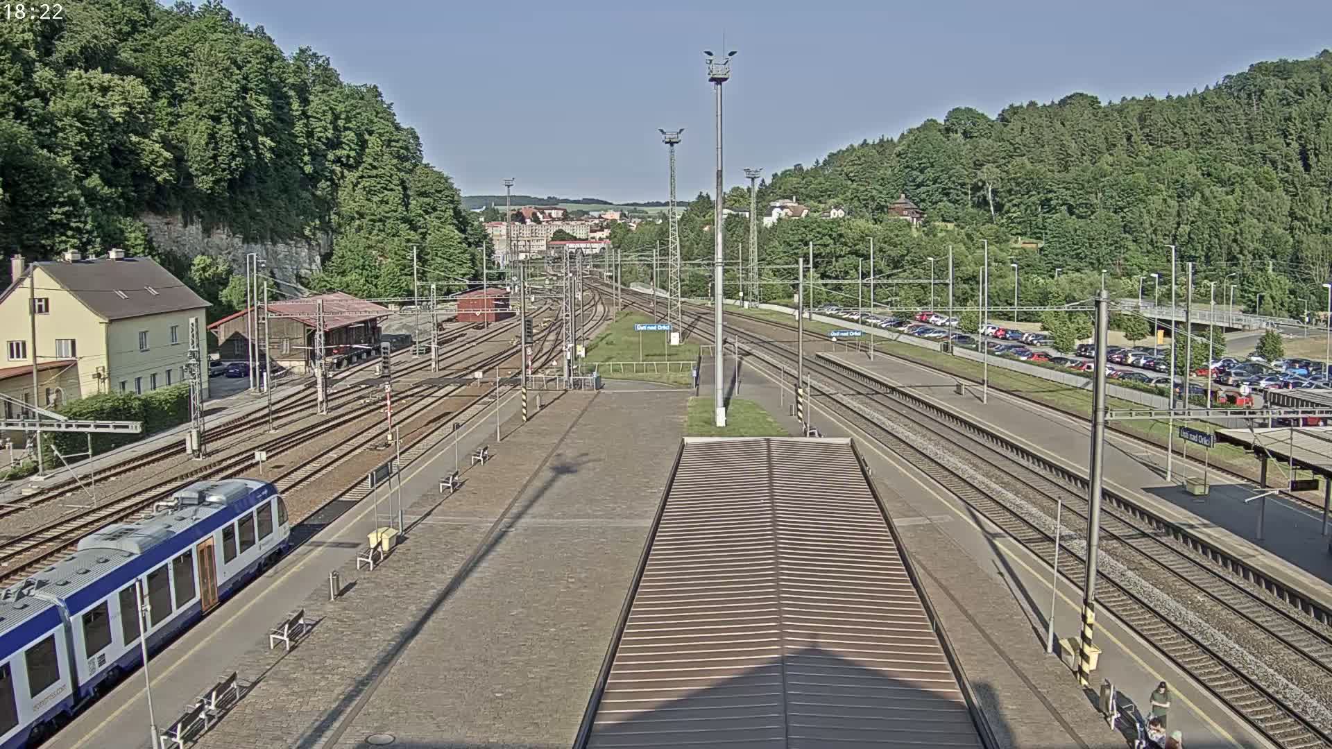 A train station with multiple train tracks, a train, and a parking lot is visible on a sunny day.