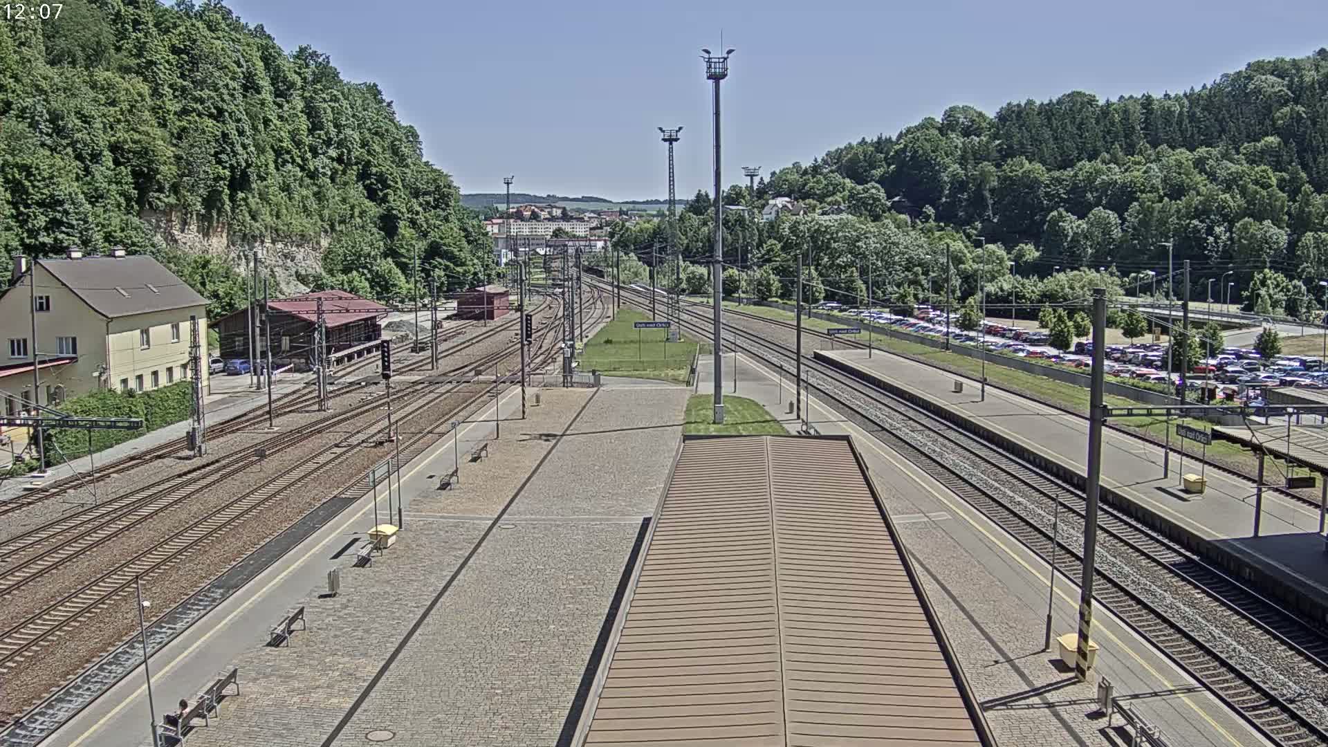 A train station on a sunny day, with multiple train tracks, a parking lot filled with cars, and lush green hills in the background.