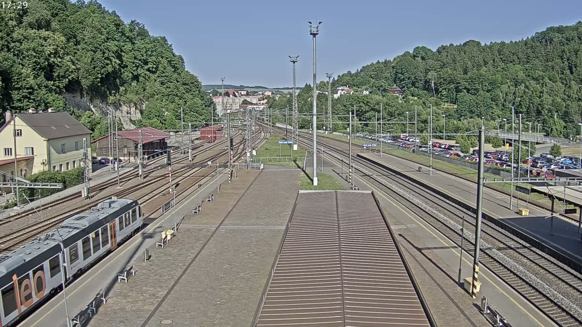 A train sits at a station on a sunny day surrounded by multiple train tracks, a parking lot, and a wooded hillside.