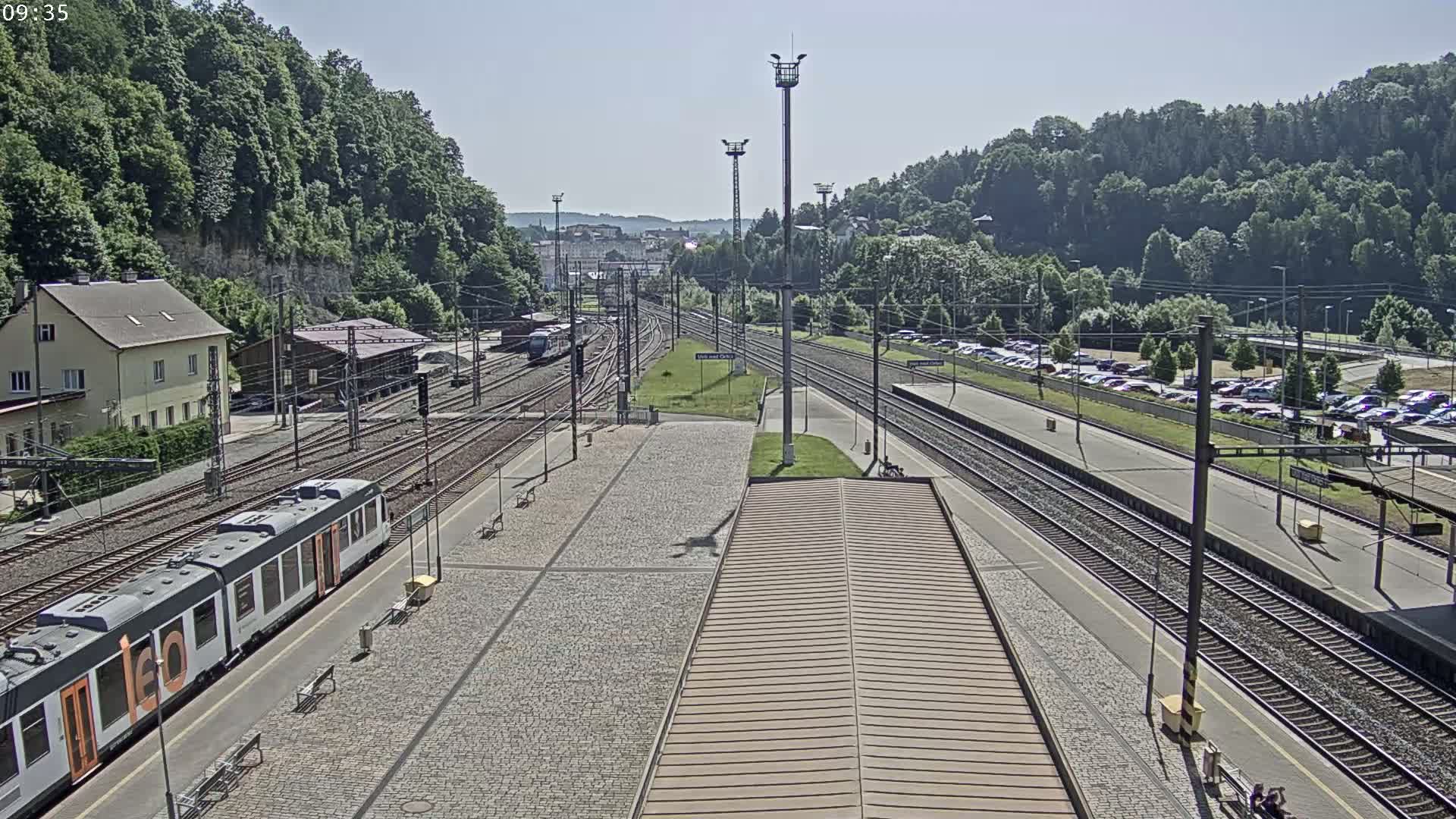 A train station on a sunny day with multiple train tracks, a train stopped at the platform, and a large parking lot visible nearby.