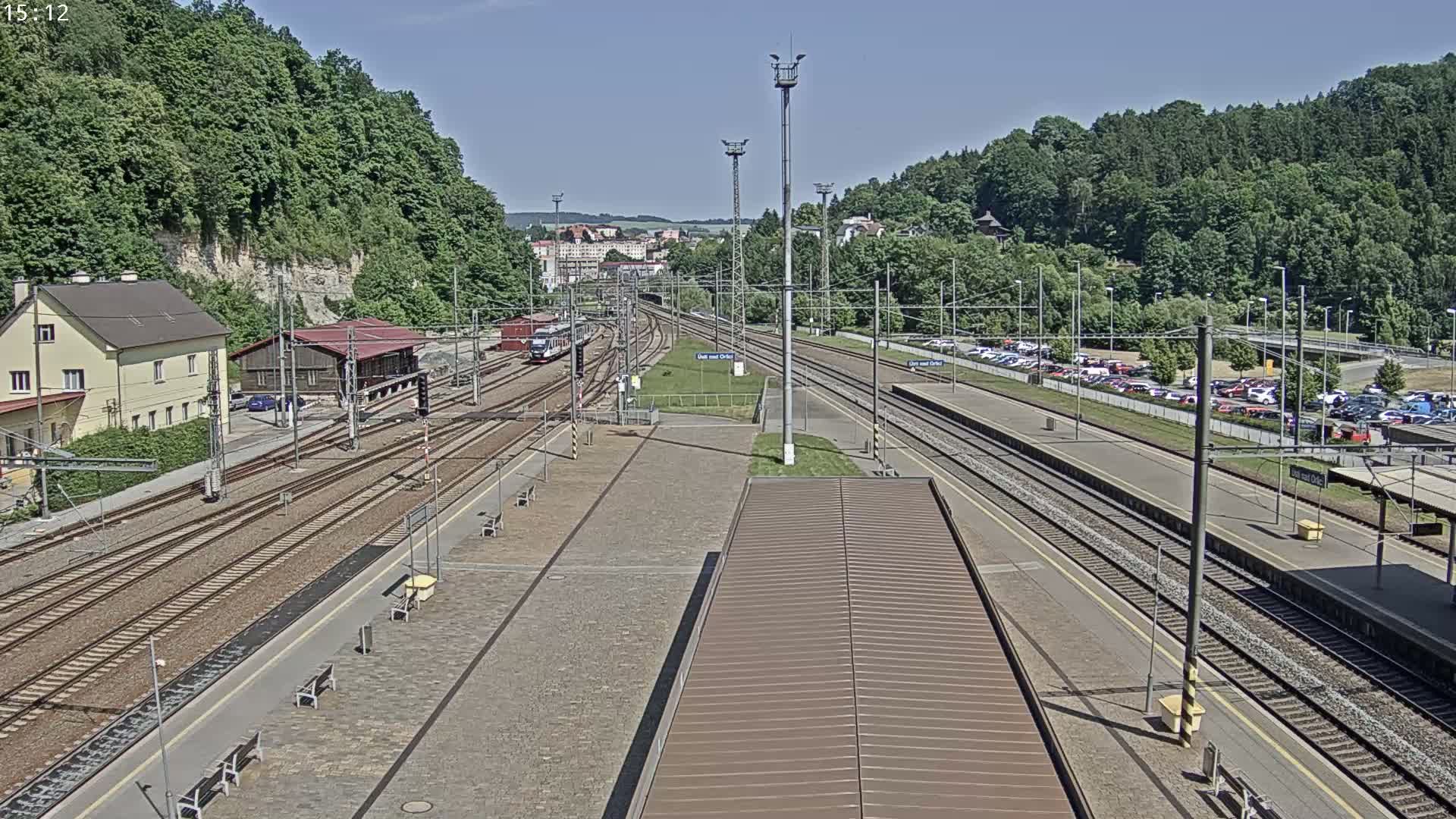 A high-angle view of a train station on a sunny day, showing multiple train tracks, platforms, a building, and a parking lot surrounded by lush green hills.