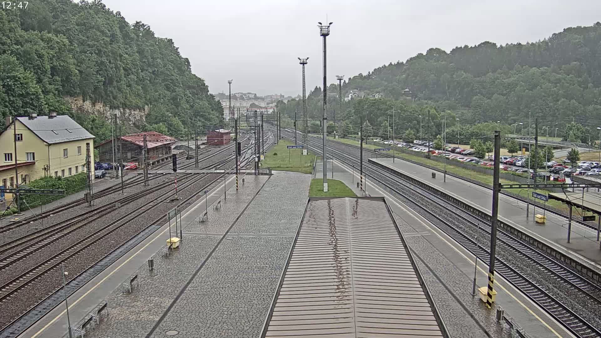 A rain-soaked train station platform with multiple train tracks, surrounded by green hills and a parking lot.