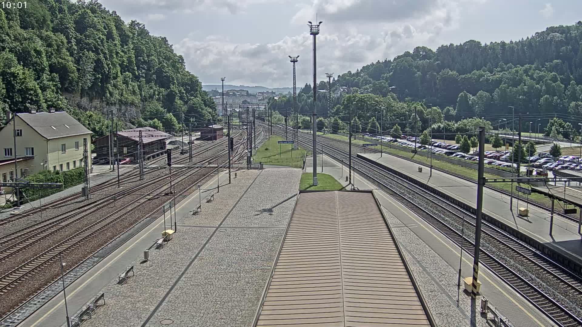 A high-angle view of a train station on a partly cloudy day, showing multiple train tracks, a platform with benches, and a large parking lot beyond the tracks, all surrounded by lush green hills.