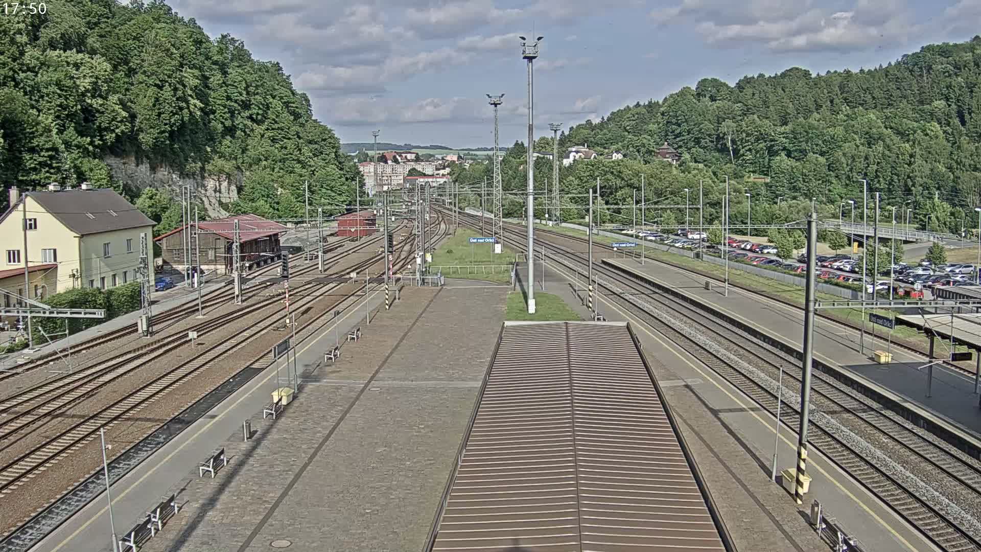 A mostly sunny day reveals a train station with numerous train tracks, platforms, and a parking lot surrounded by lush green hills.