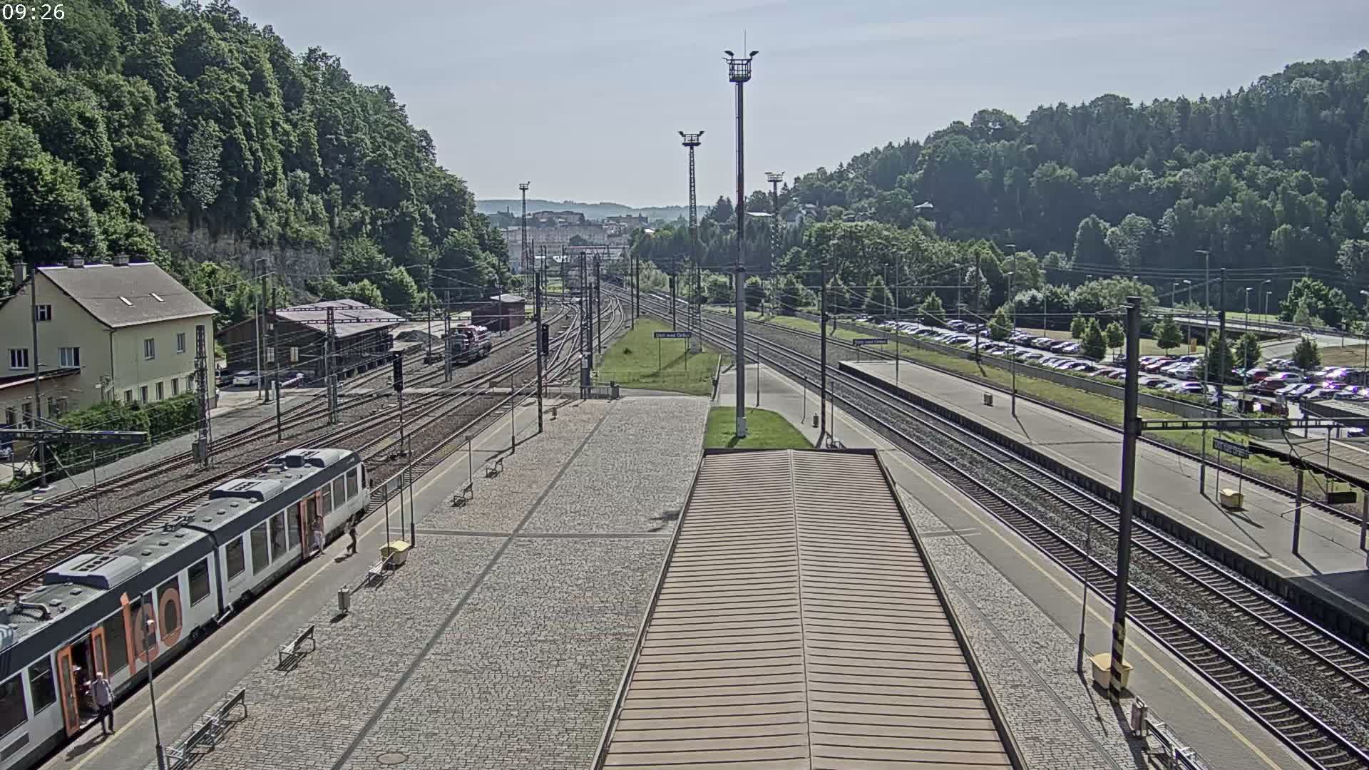 A train station on a sunny day with a train at the platform, multiple tracks, and a large parking lot visible beyond the tracks.