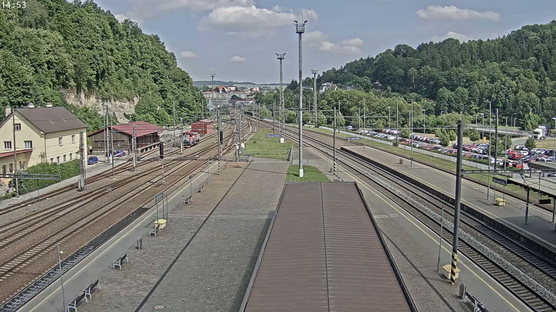A high-angle view of a train station on a partly cloudy day, showing multiple train tracks, platforms, a building, and a large parking lot.