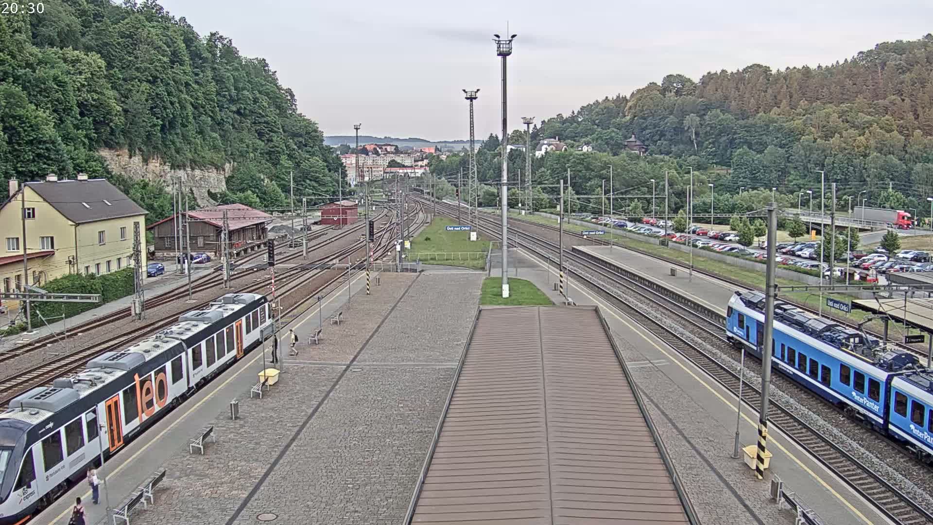 A high-angle view of a train station on a partly cloudy day, showing multiple train tracks, two passenger trains, and a parking lot filled with cars.