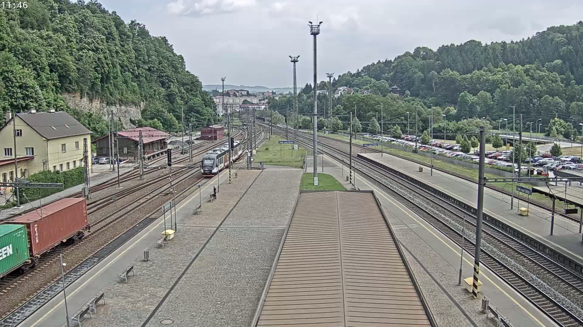 A train is arriving at a train station on an overcast day, with lush green hills in the background and several cars parked in lots nearby.
