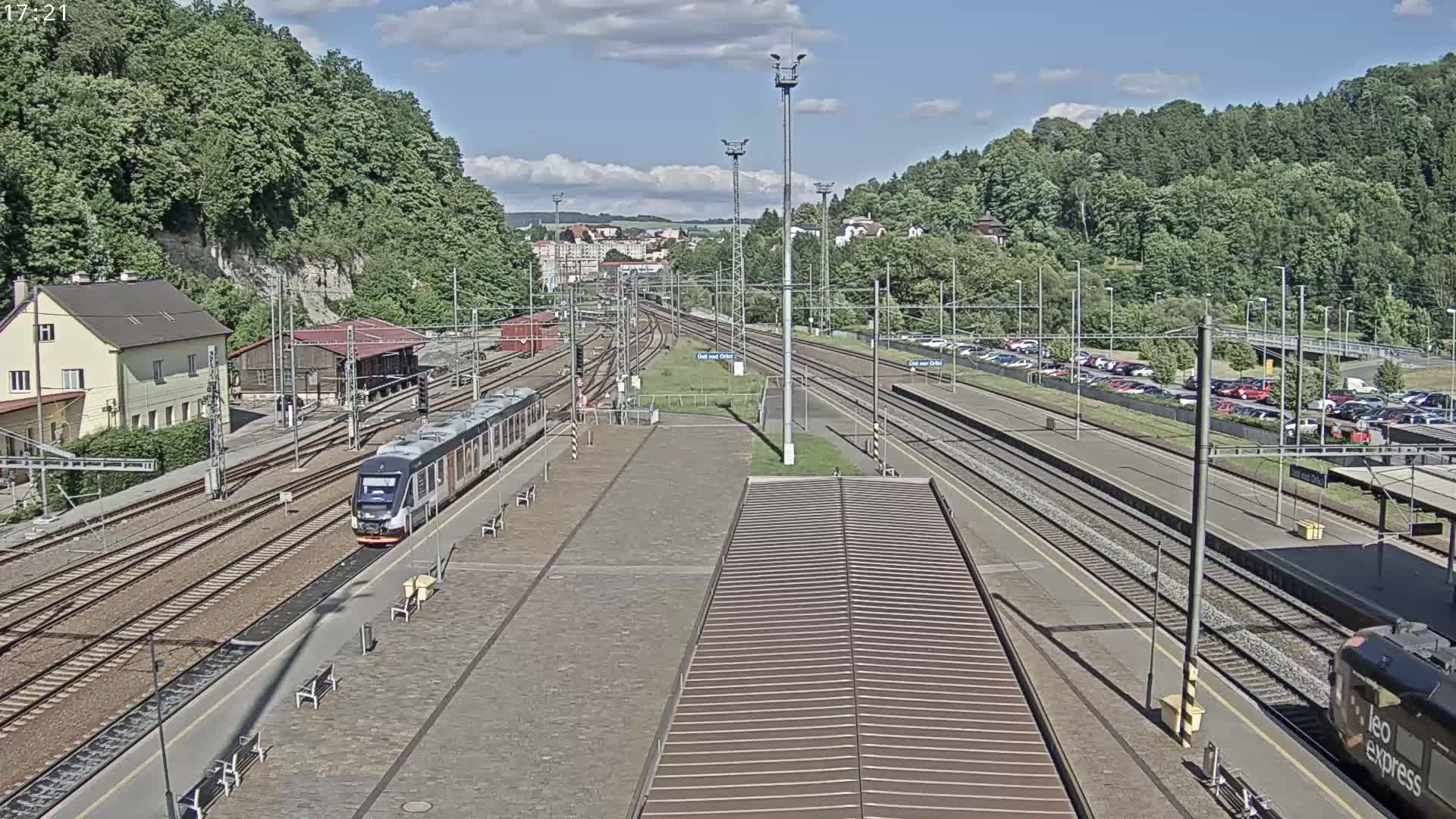 A train arrives at a railway station on a partly cloudy day, with hills covered in lush green trees in the background.