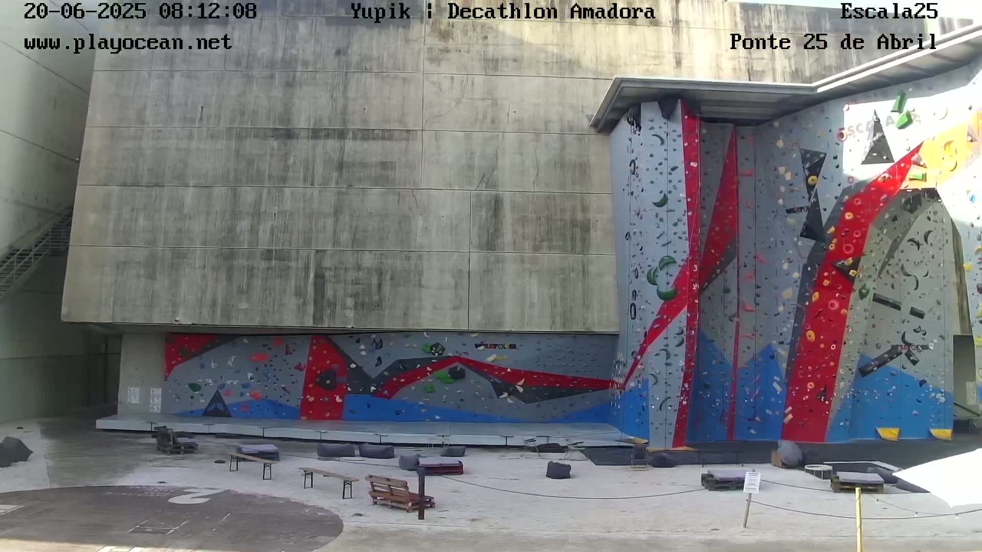 A large outdoor climbing wall with various colored holds is situated against a concrete building on a sunny day.