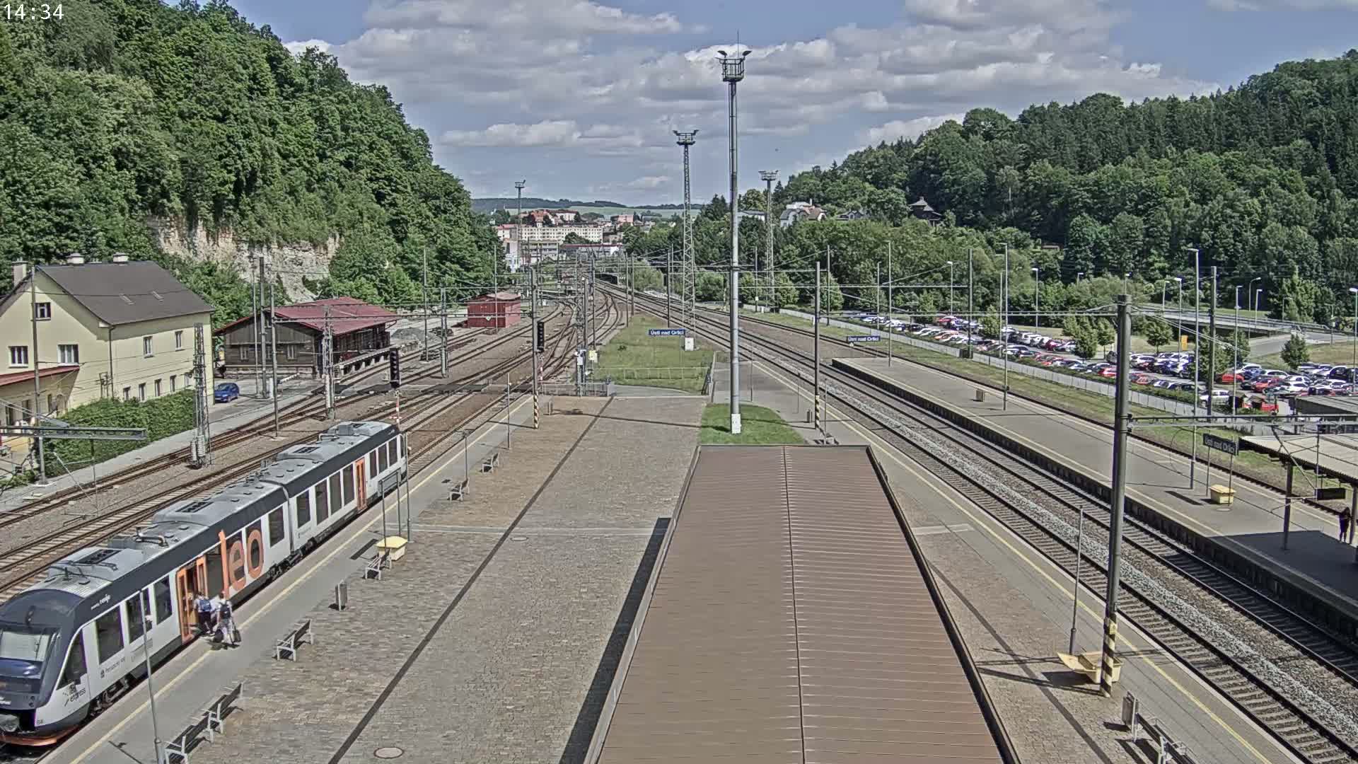 A train sits at a station platform next to several railway tracks, under a partly cloudy sky, with a large parking lot and lush green hills in the background.