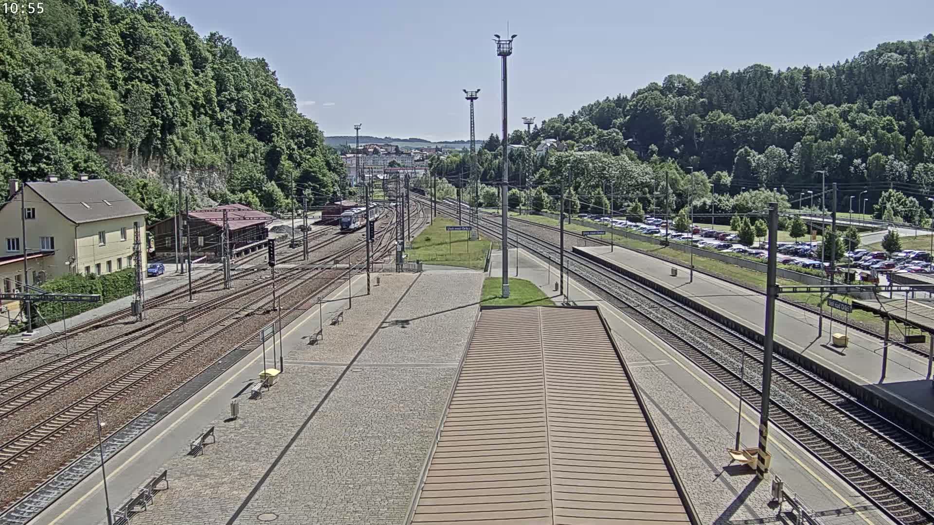 A train station on a sunny day with multiple train tracks, a large parking lot, and lush green hills in the background.