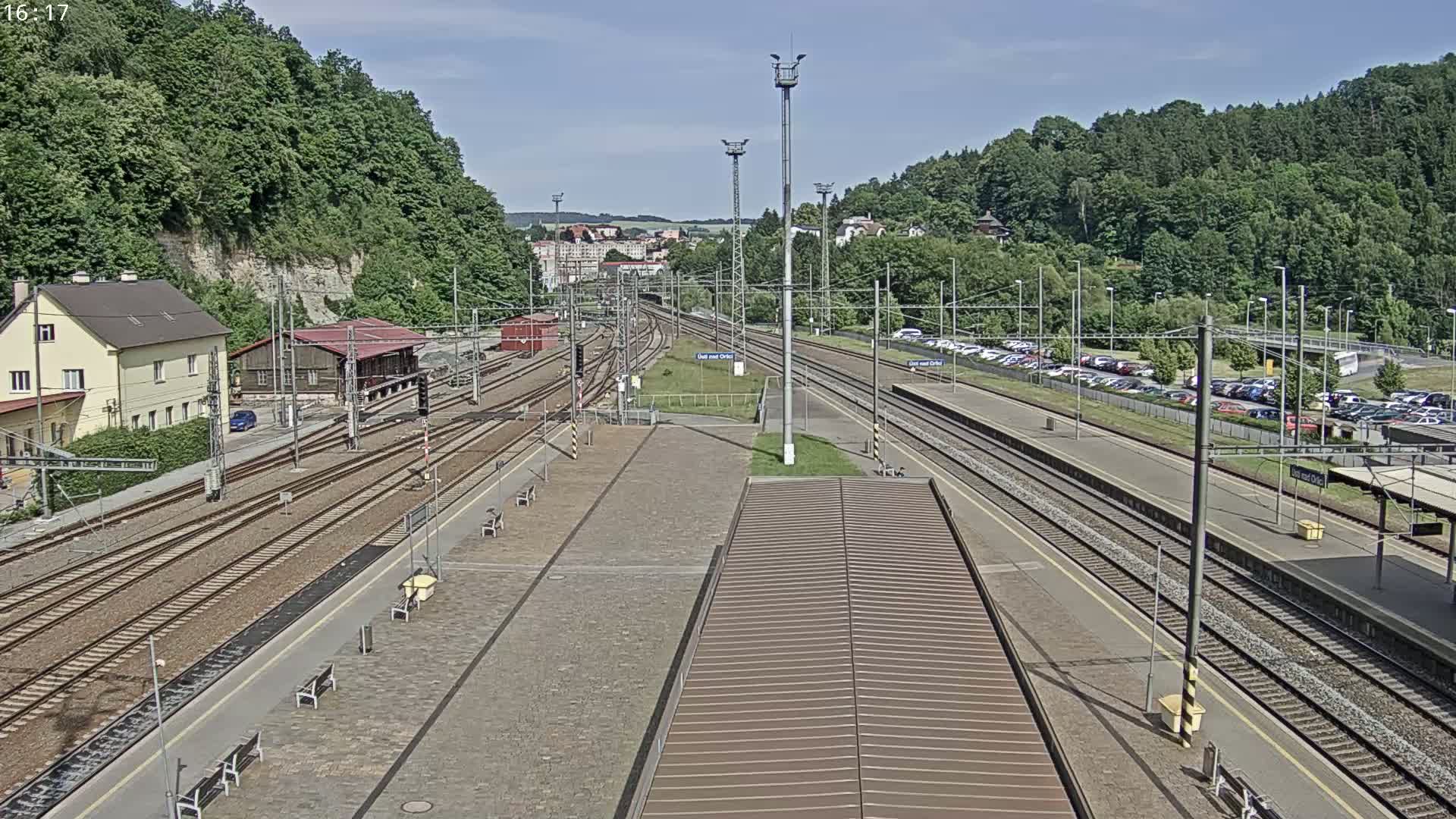 A sunny day view of a train station with multiple tracks, platforms, and a large parking lot, surrounded by lush green hills.
