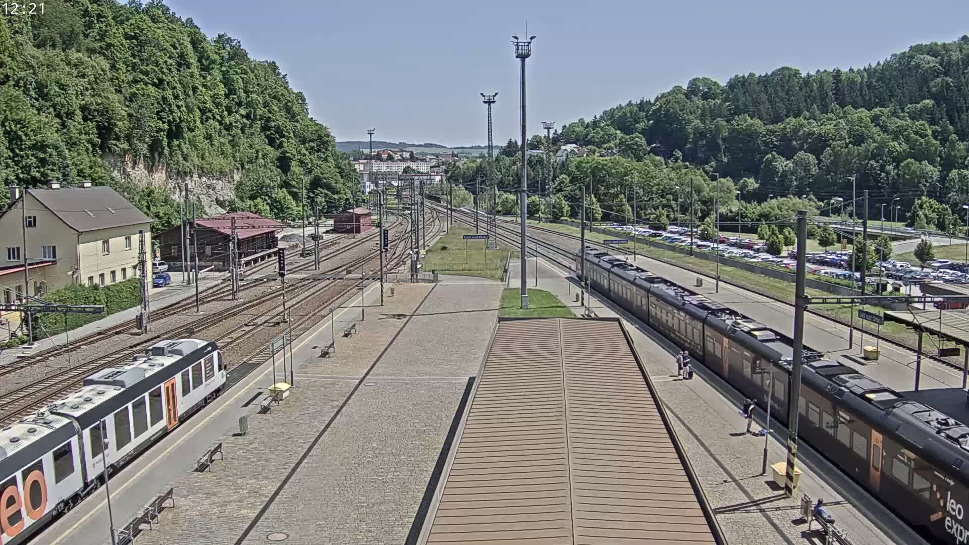 A train station on a sunny day with two trains at the platform, surrounded by lush green hills and a parking lot.