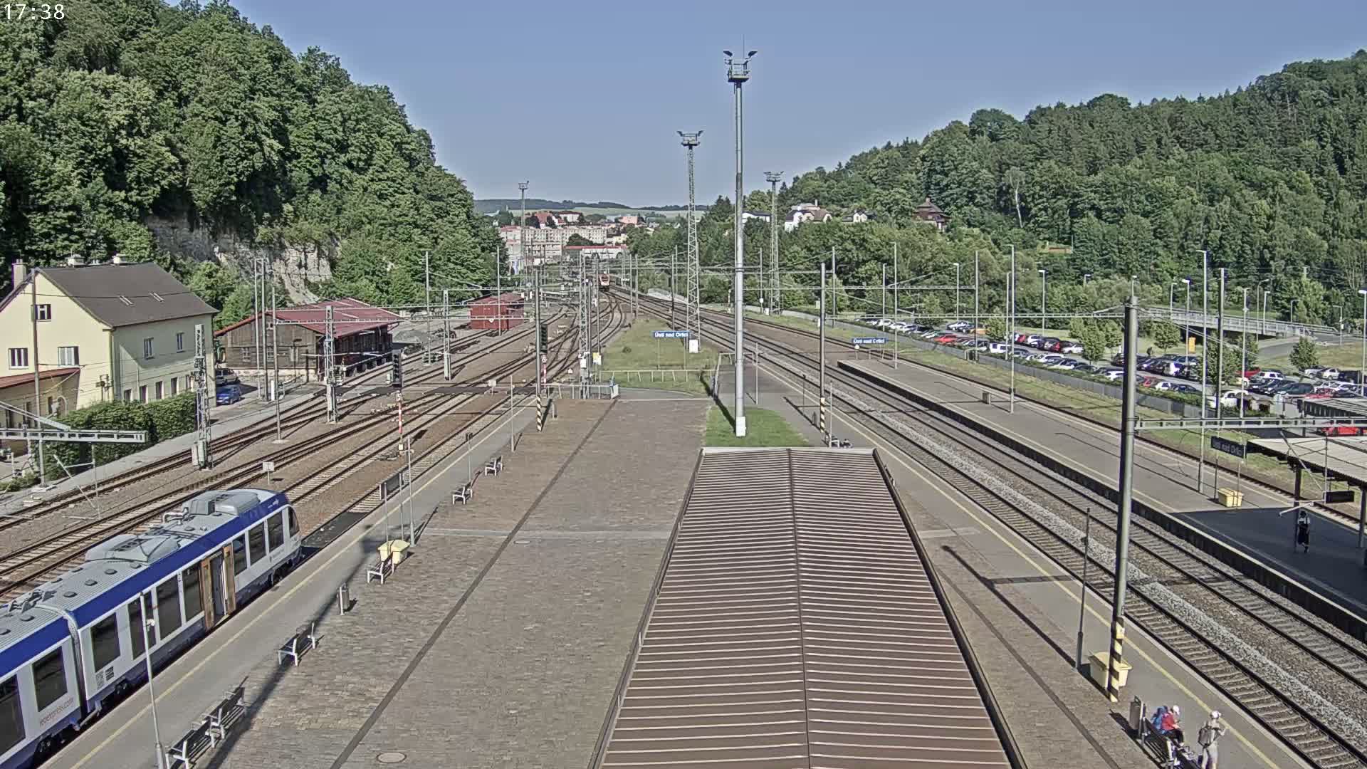 A train station with multiple tracks, a train, and a parking lot is visible on a sunny day, surrounded by lush green hills.
