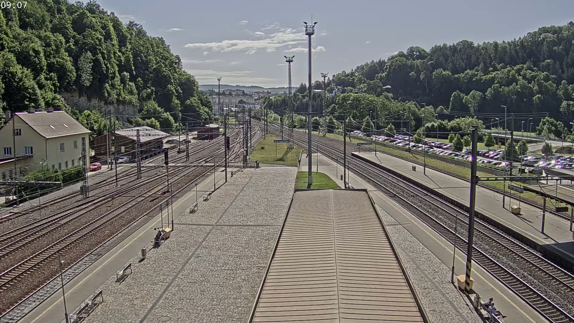 A high-angle view of a train station on a sunny day, showing multiple train tracks, platforms, a parking lot, and a surrounding landscape of green hills.