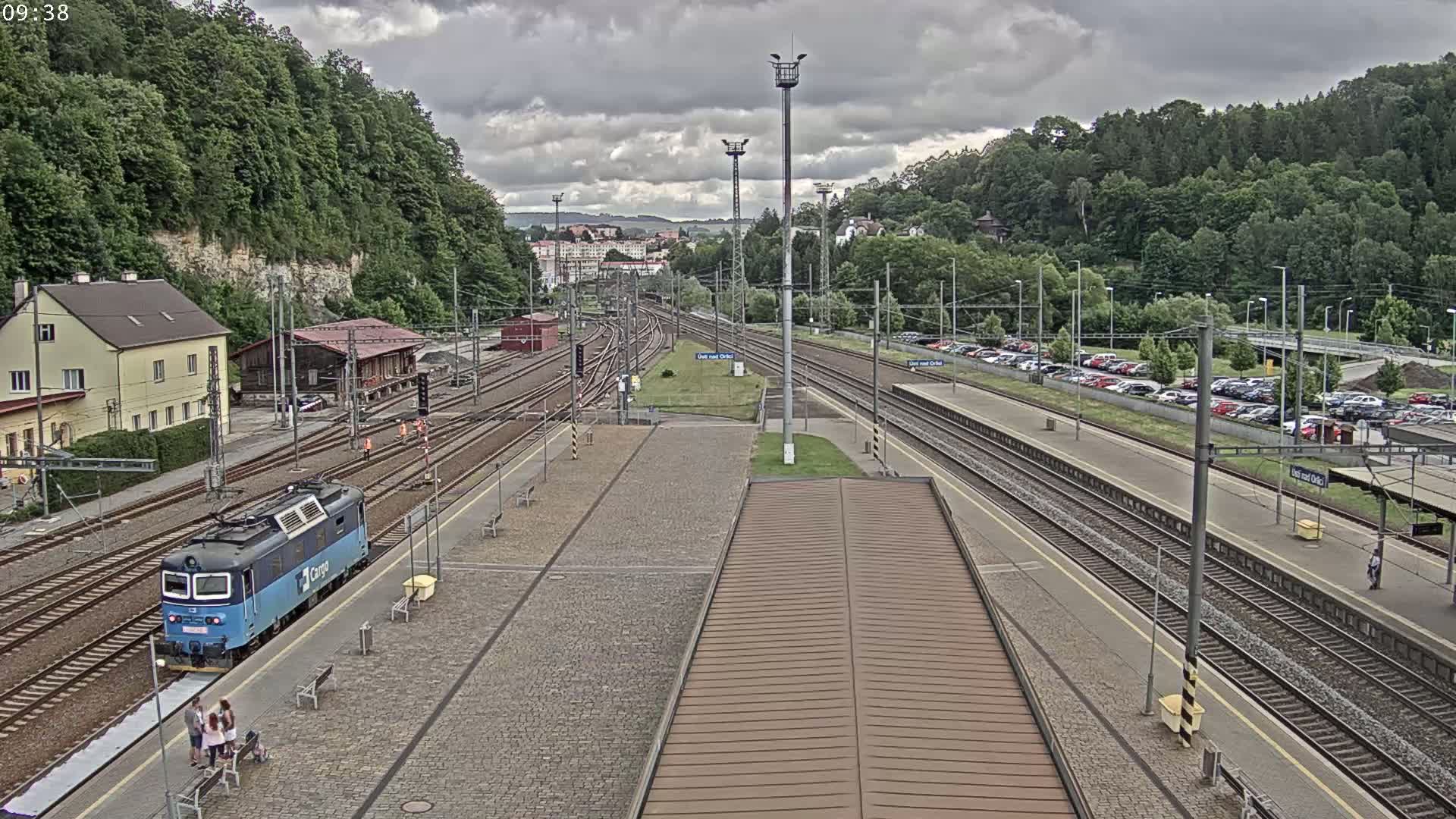 A train station with multiple tracks, a blue locomotive, a parking lot, and green hills under a cloudy sky.
