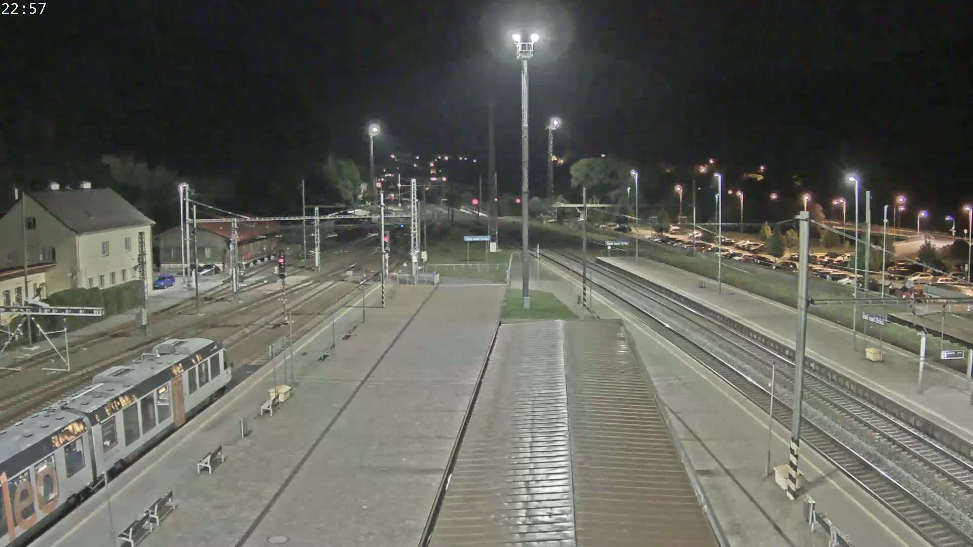 A nighttime view of a train station with a train at a platform, multiple tracks, and a parking lot visible in the background under dark skies.