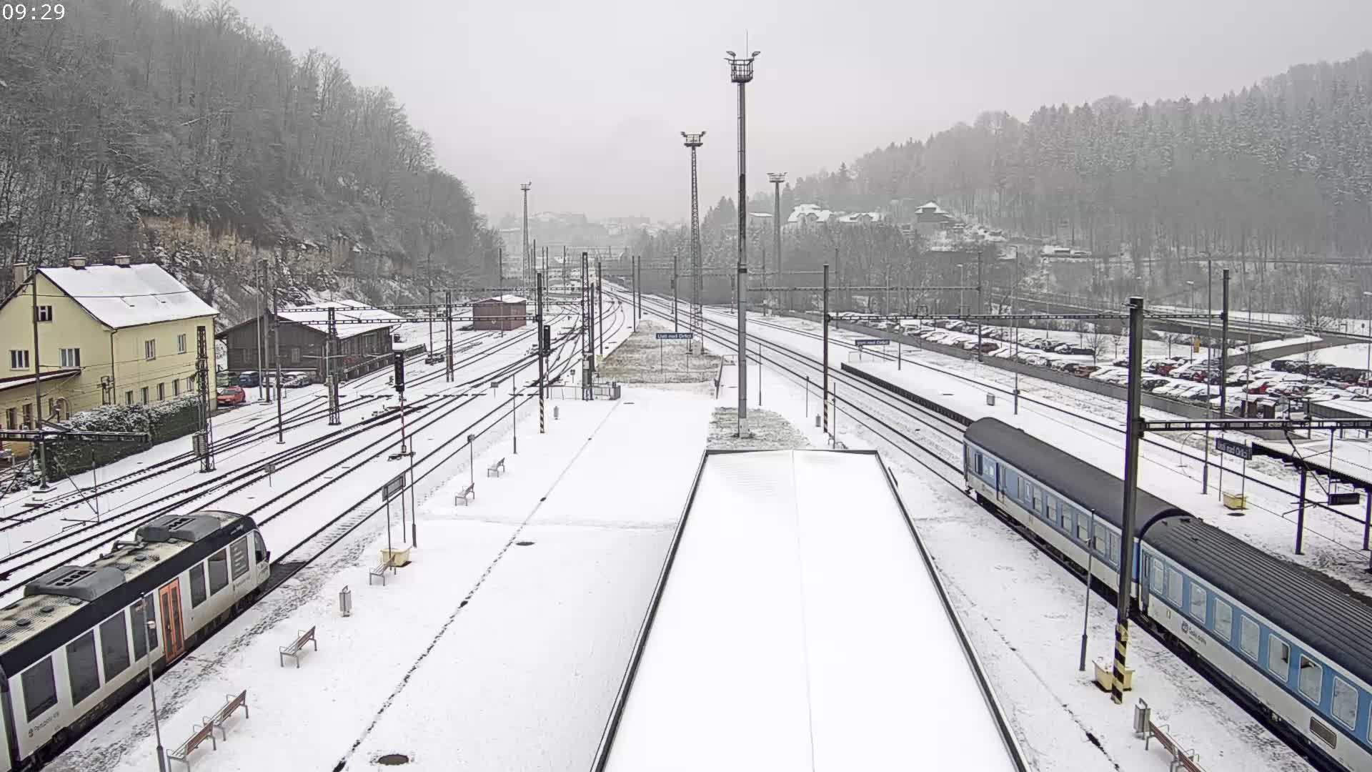 A wide view of a snow-covered train station reveals multiple tracks, two trains at platforms, various buildings, a parking lot full of cars, and distant forested hills, all under a dim, cloudy winter sky.