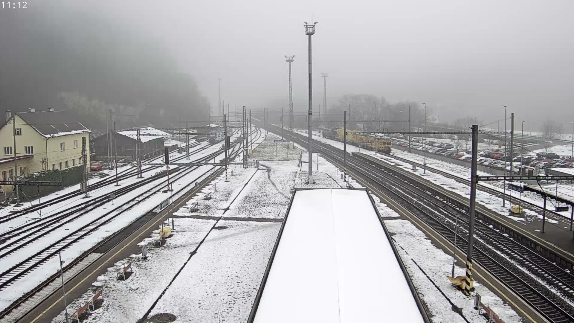 A snow-covered railway yard with numerous tracks, platforms, and adjacent buildings, alongside a car park, all stretch into the dense fog.