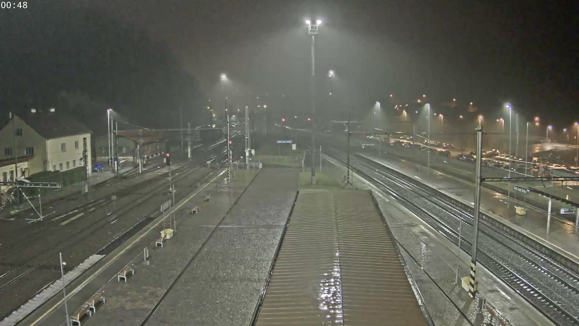 The image displays a multi-track railway station at night, with wet platforms and tracks glistening under numerous streetlights as rain visibly falls through the misty air.