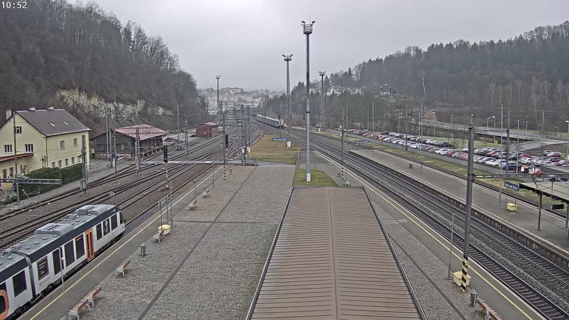 The image displays a multi-track railway station at night, with wet platforms and tracks glistening under numerous streetlights as rain visibly falls through the misty air.