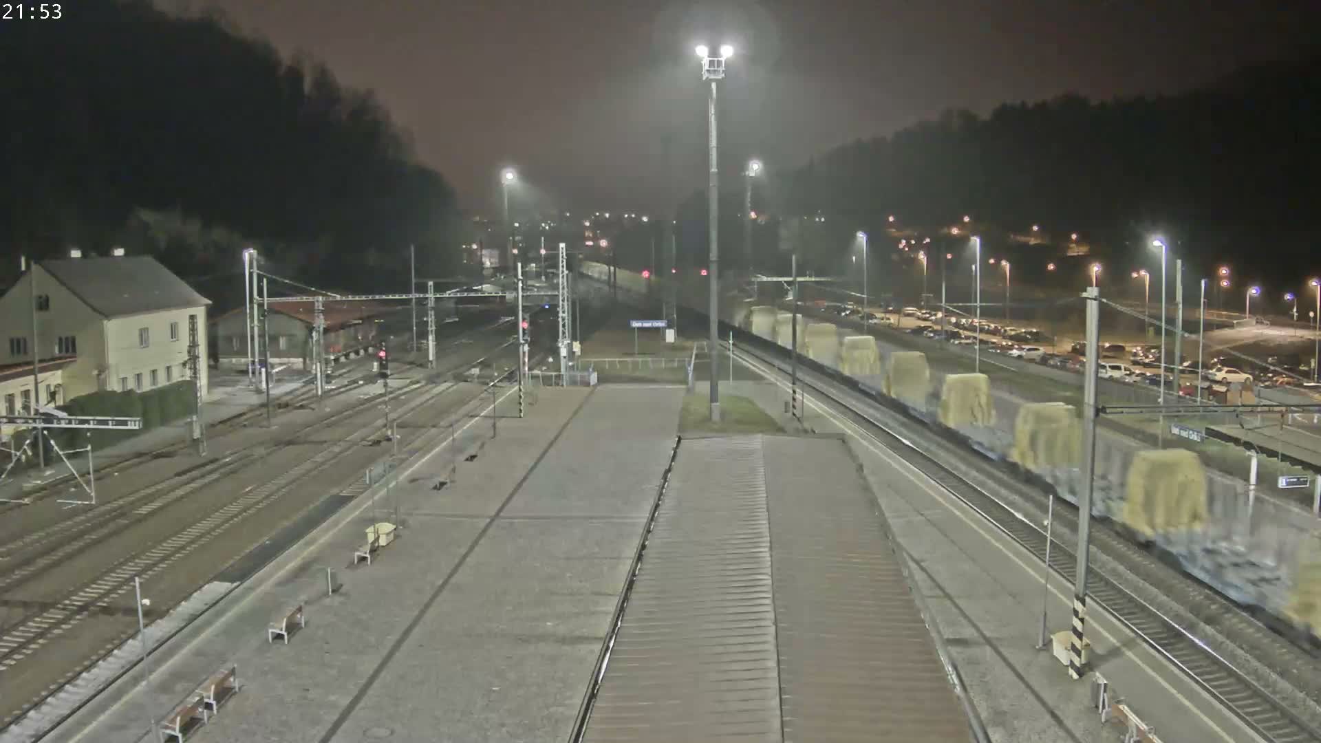 The image displays a multi-track railway station at night, with wet platforms and tracks glistening under numerous streetlights as rain visibly falls through the misty air.