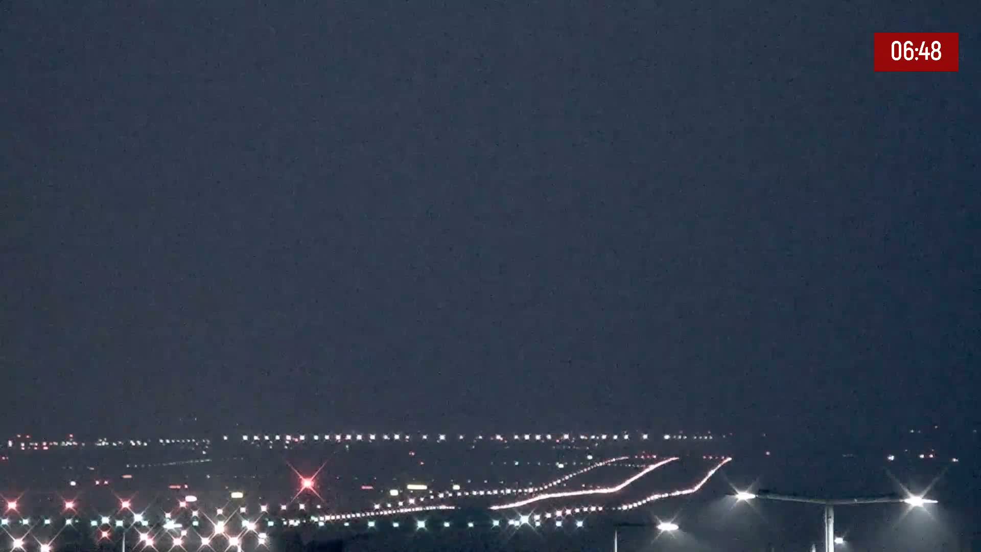 Under a dark night sky, the lights of a distant city and airport runway are visible, with illuminated streetlights in the foreground.